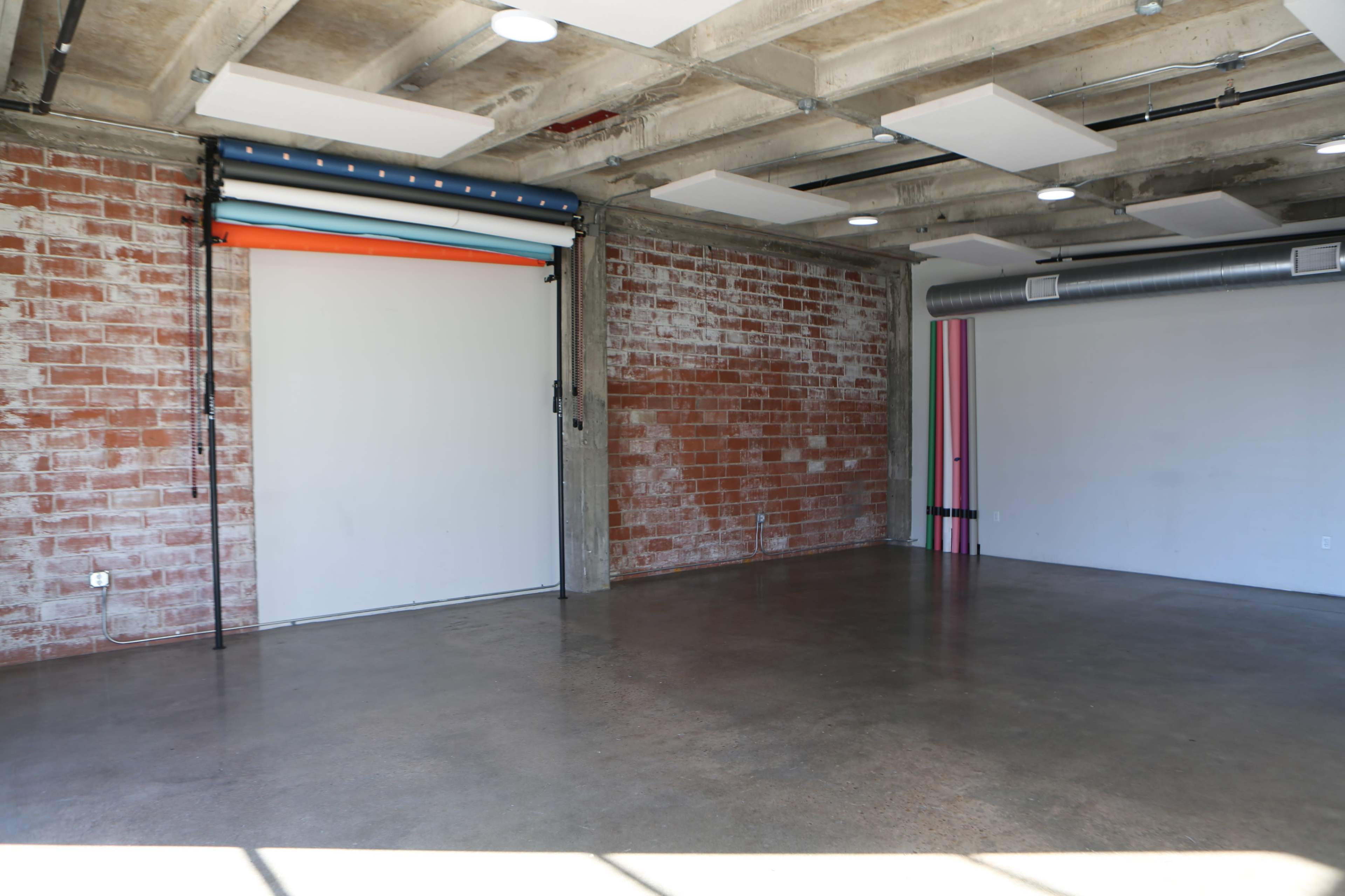 The image shows an industrial-style studio with exposed brick walls, a concrete floor, and a large sliding door with colorful backdrop rolls.
