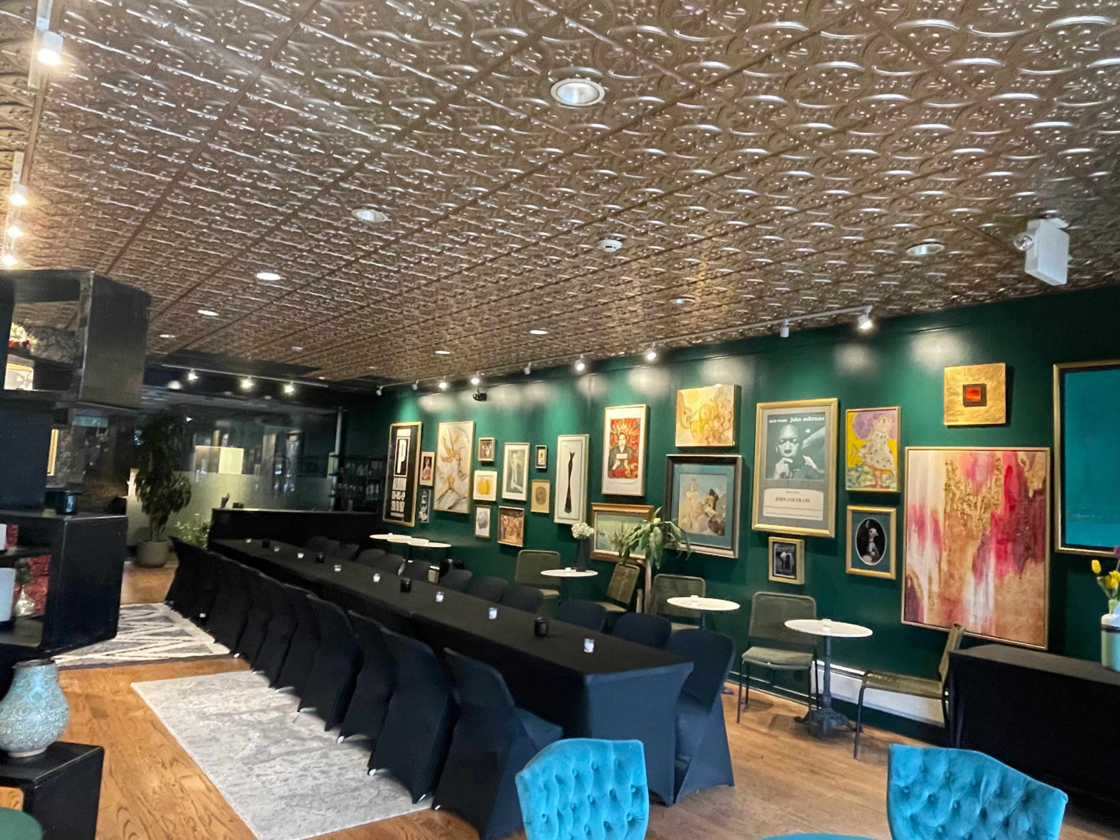 The image shows a decorated event space with a long black table set for a gathering, surrounded by various artworks on the green walls, and featuring a patterned ceiling.