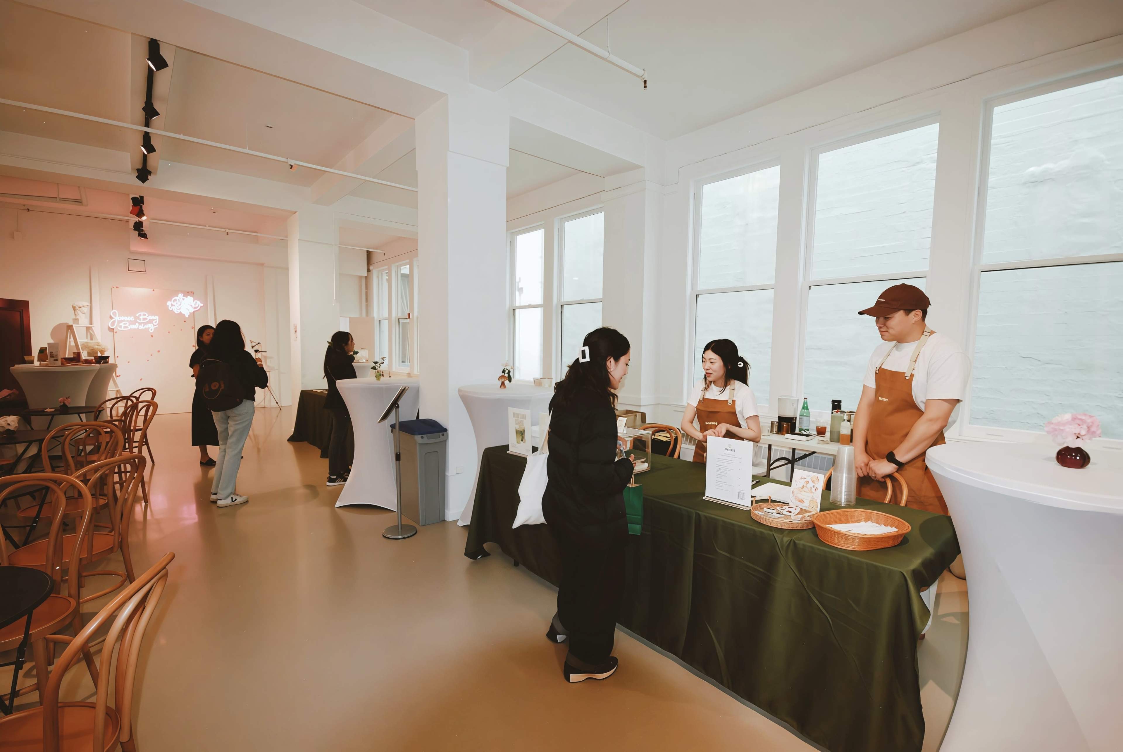 A modern café interior features a green counter where staff serve customers, with tables and chairs arranged throughout the bright space.