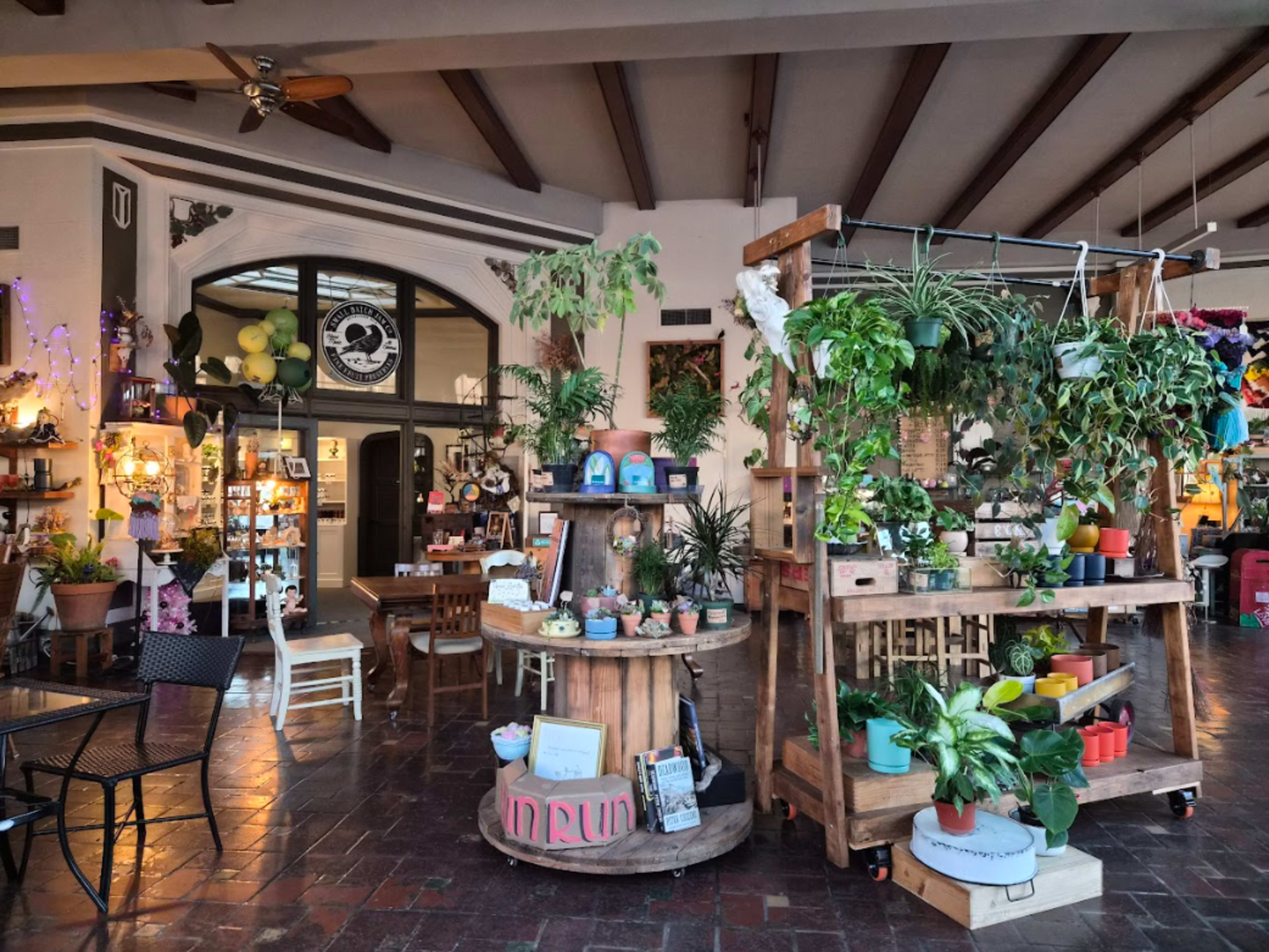 The image shows a cozy indoor market with wooden displays filled with various potted plants and decorations, alongside a seating area with black tables and chairs.