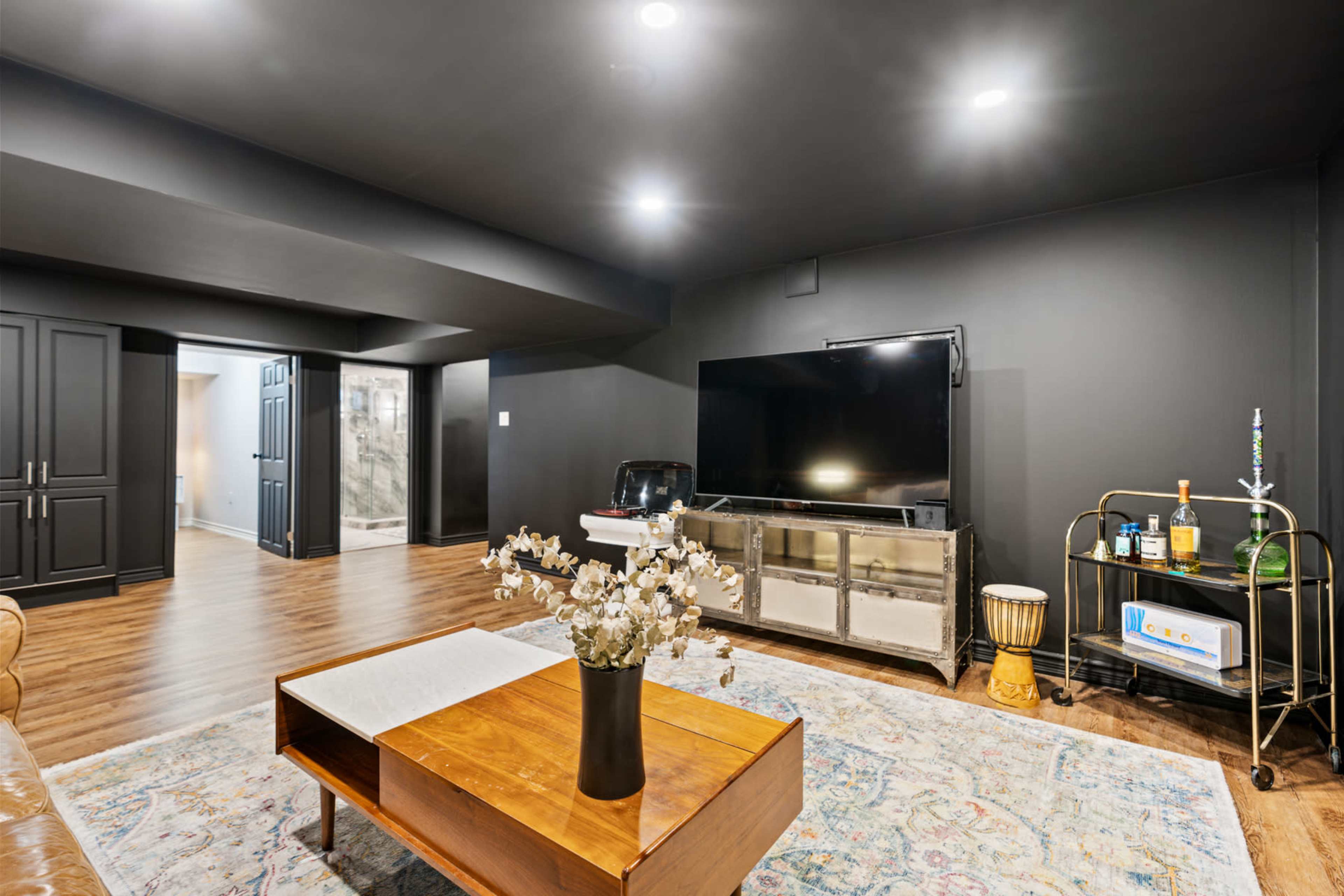 A modern basement living area features a large television mounted on a dark wall, a wooden coffee table with a vase of flowers, and a bar cart with beverages.