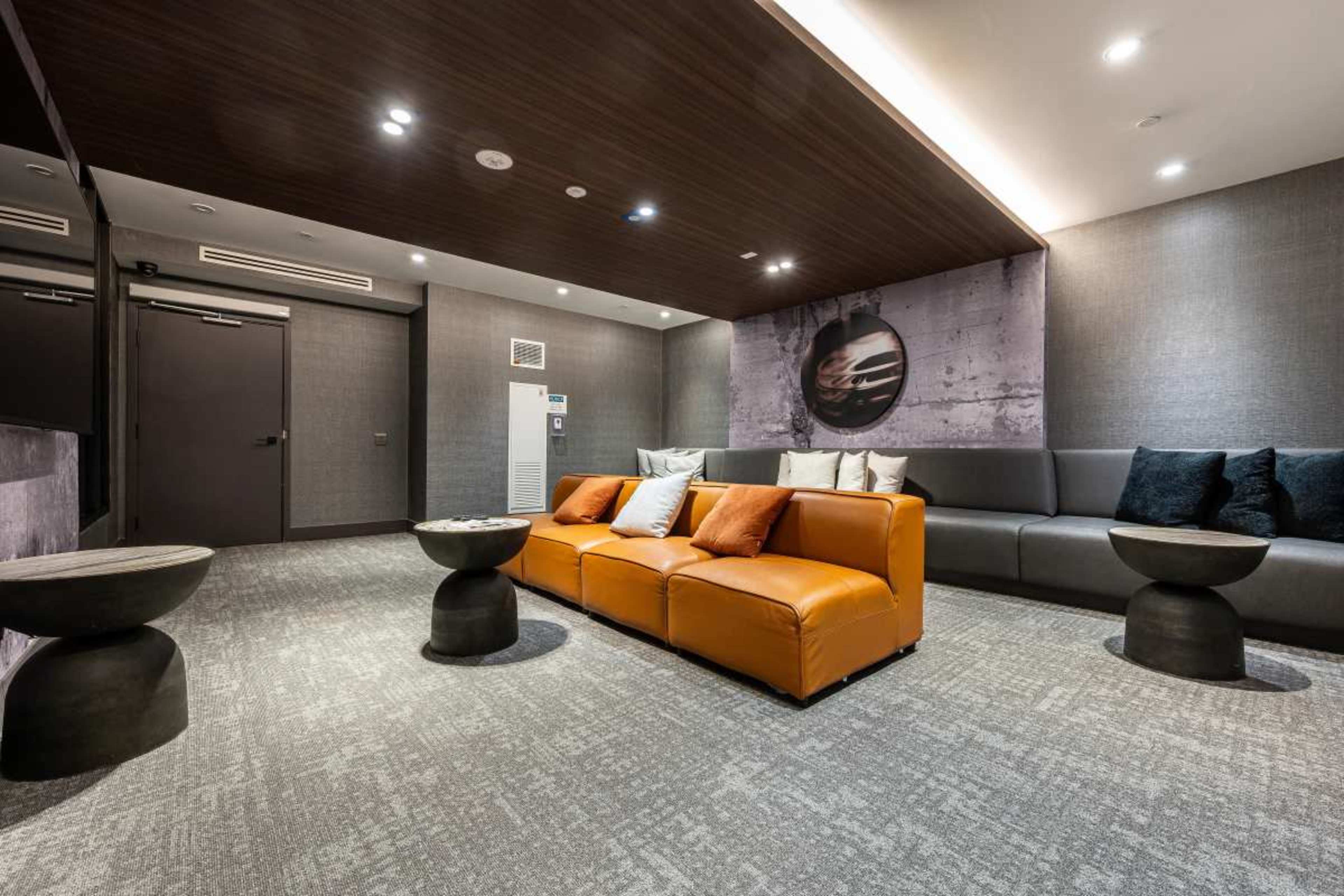 The image shows a modern lounge area featuring a large orange sofa, several decorative pillows, and black side tables on a carpeted floor.