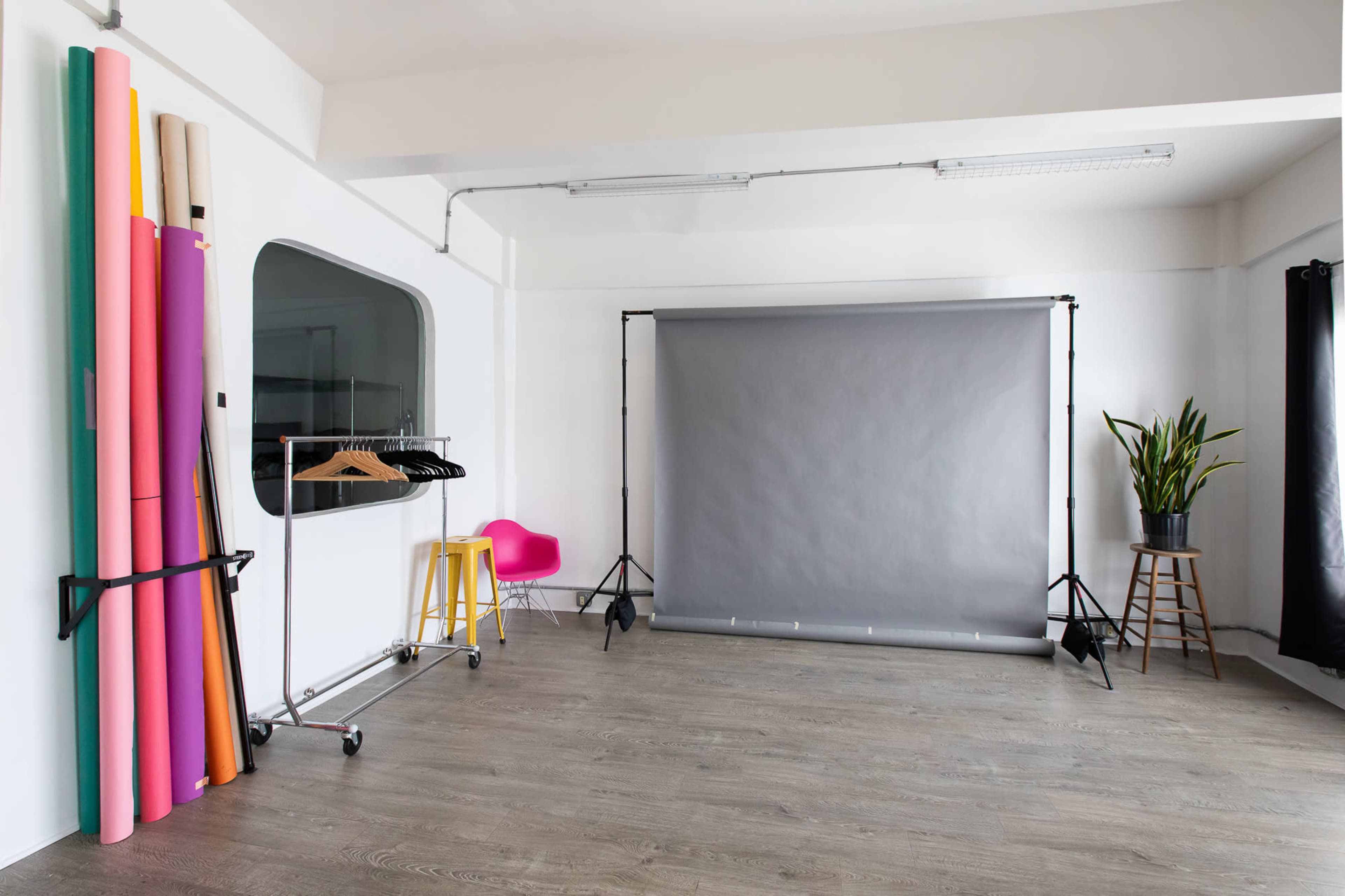 The image shows a photography studio featuring a gray backdrop, colorful rolls of paper, clothing on a rack, and potted plants.