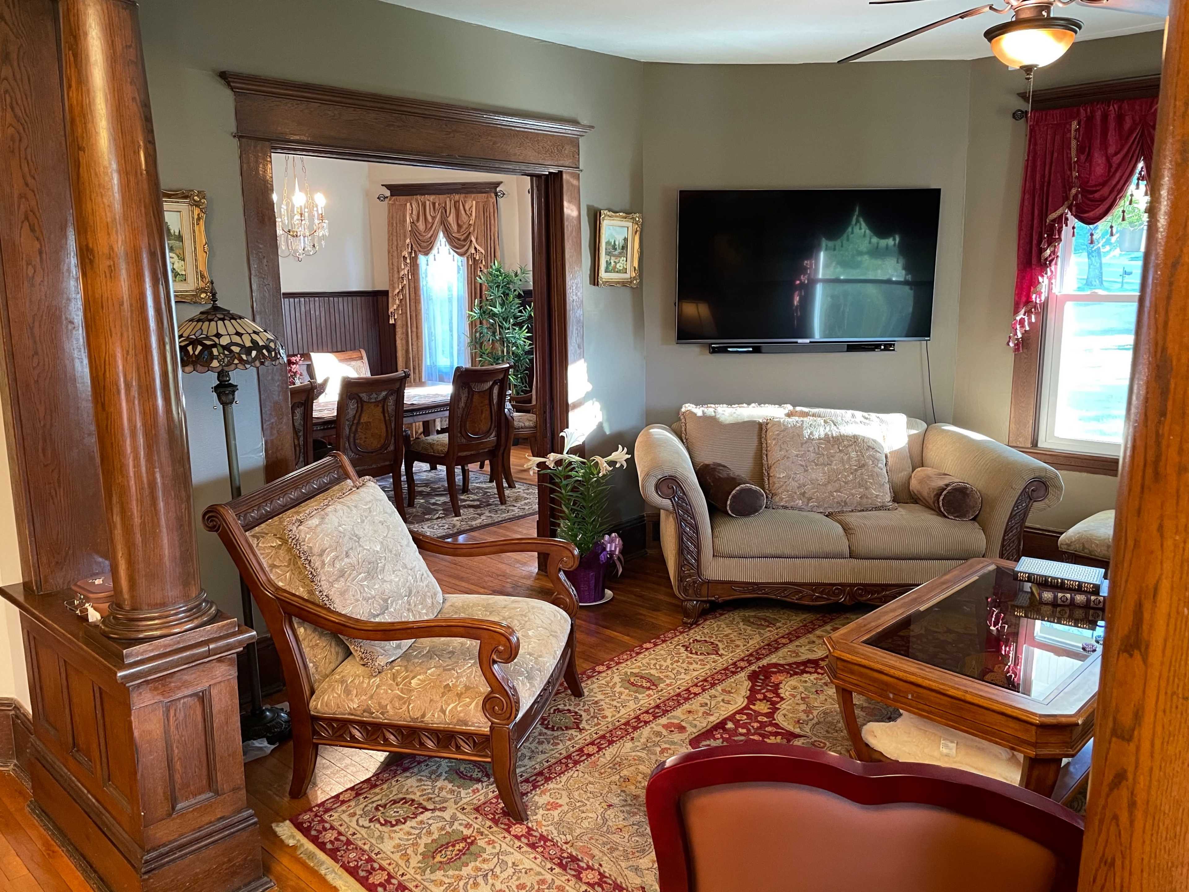 A cozy living room with a beige sofa, a wooden chair, and a TV mounted on the wall, leading to a formal dining area in the background.