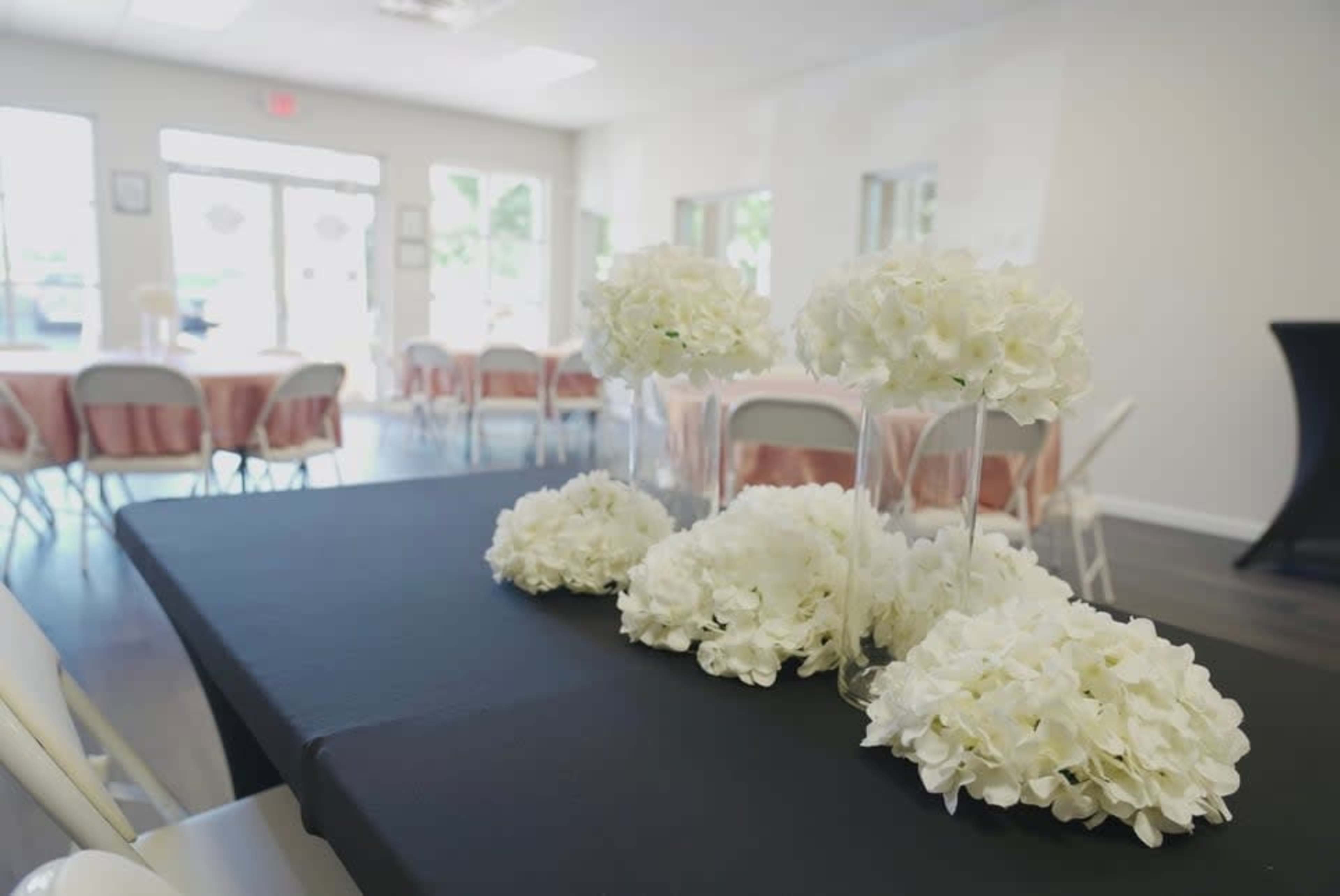 A black table centerpiece features white hydrangeas, surrounded by several tables set for an event in a well-lit room.