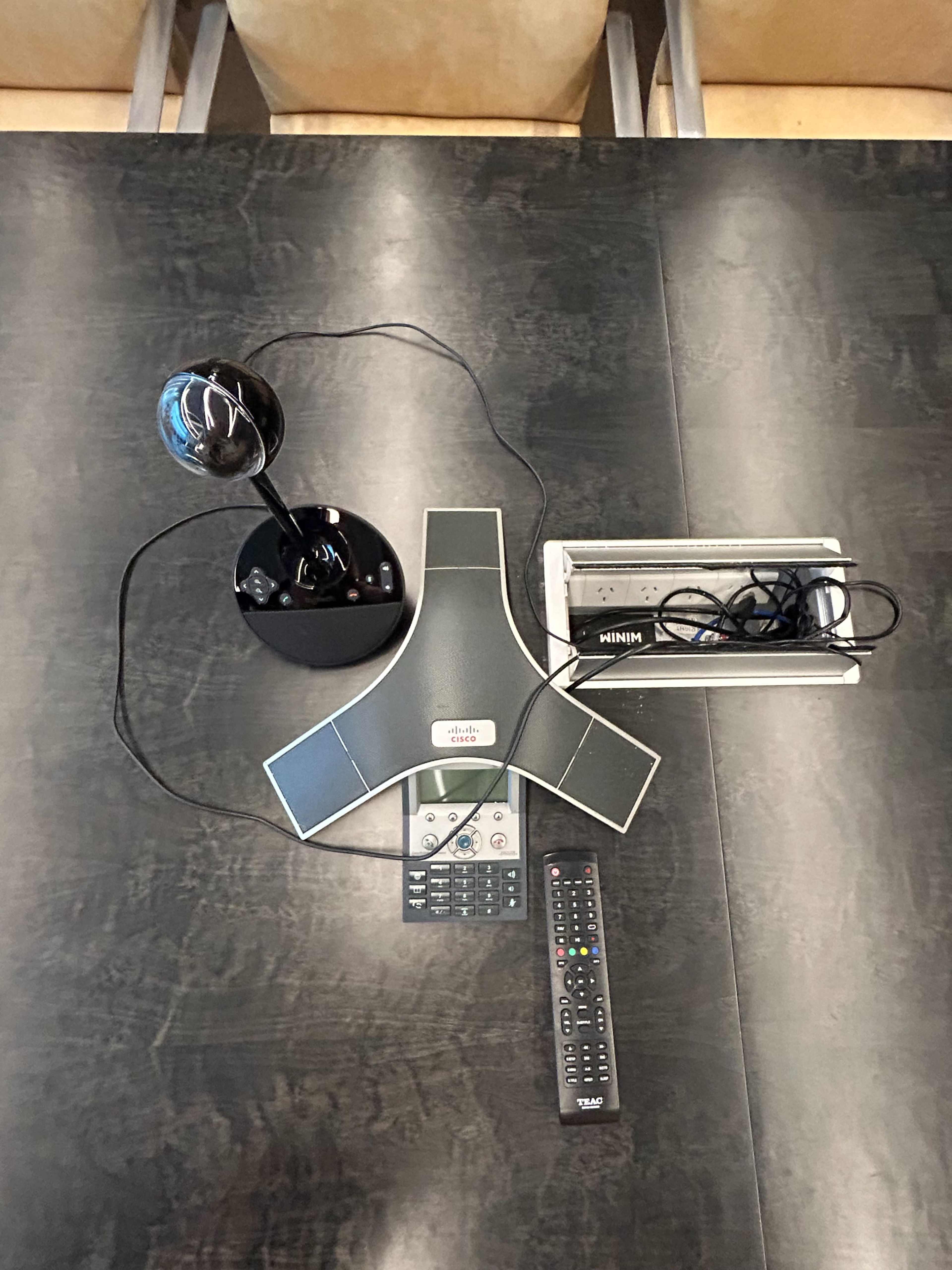 The image shows a conference phone, a microphone on a stand, a remote control, and a power strip arranged on a dark table.