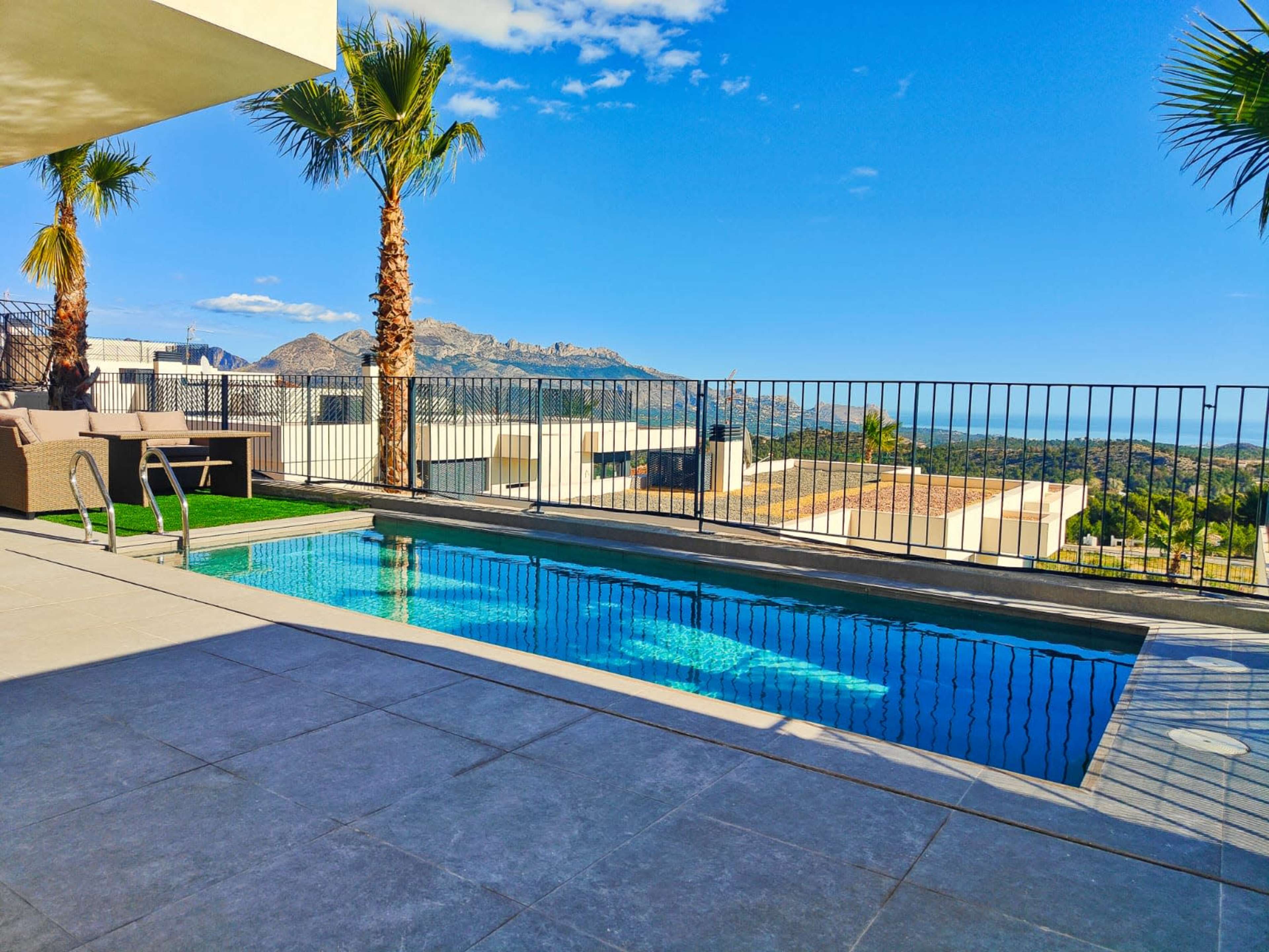 The image shows a modern swimming pool surrounded by a patio with palm trees, overlooking a mountainous landscape under a clear blue sky.