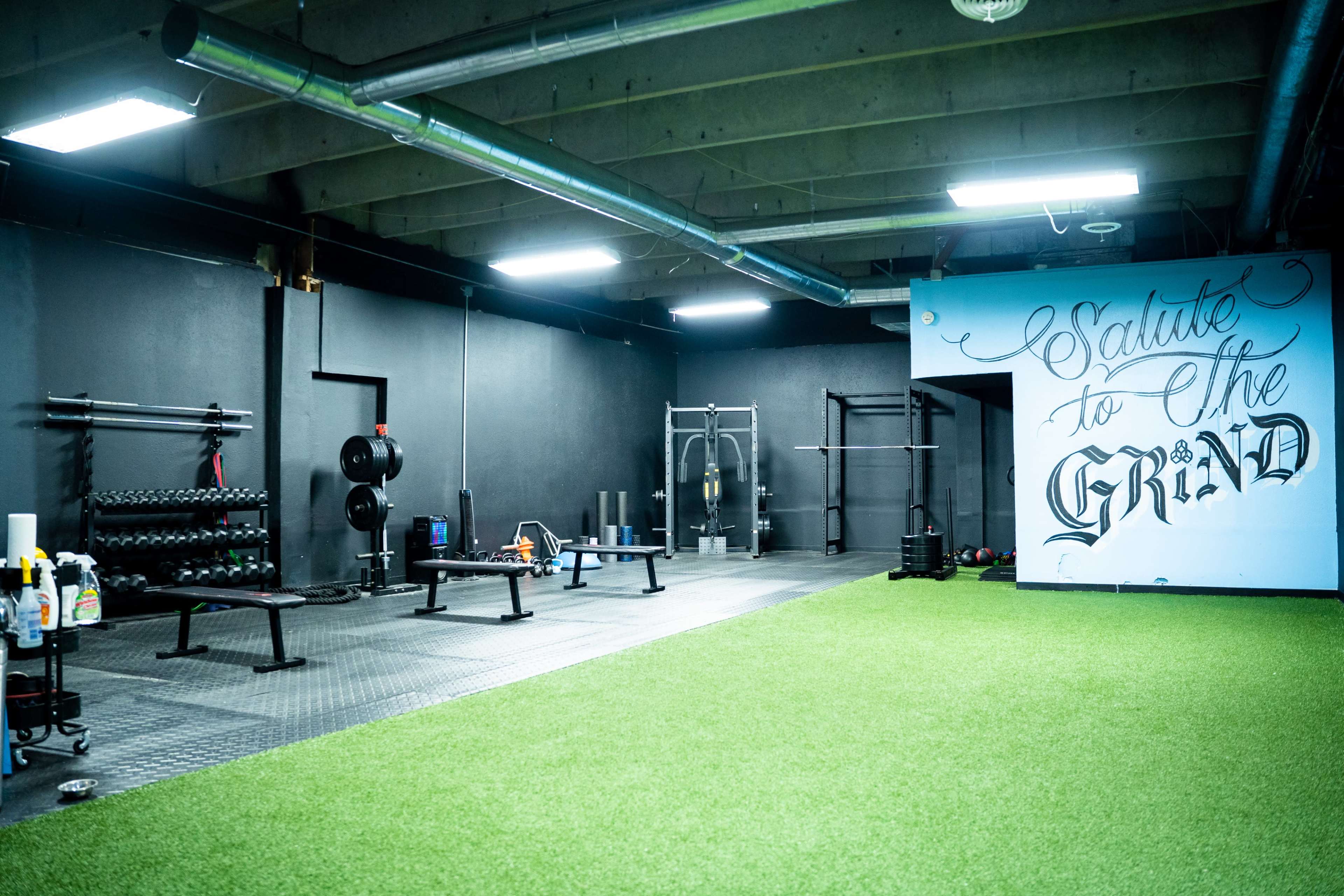 A fitness studio features various weightlifting equipment and a turf area, with one wall adorned with a mural that reads "Salute to the Grind."