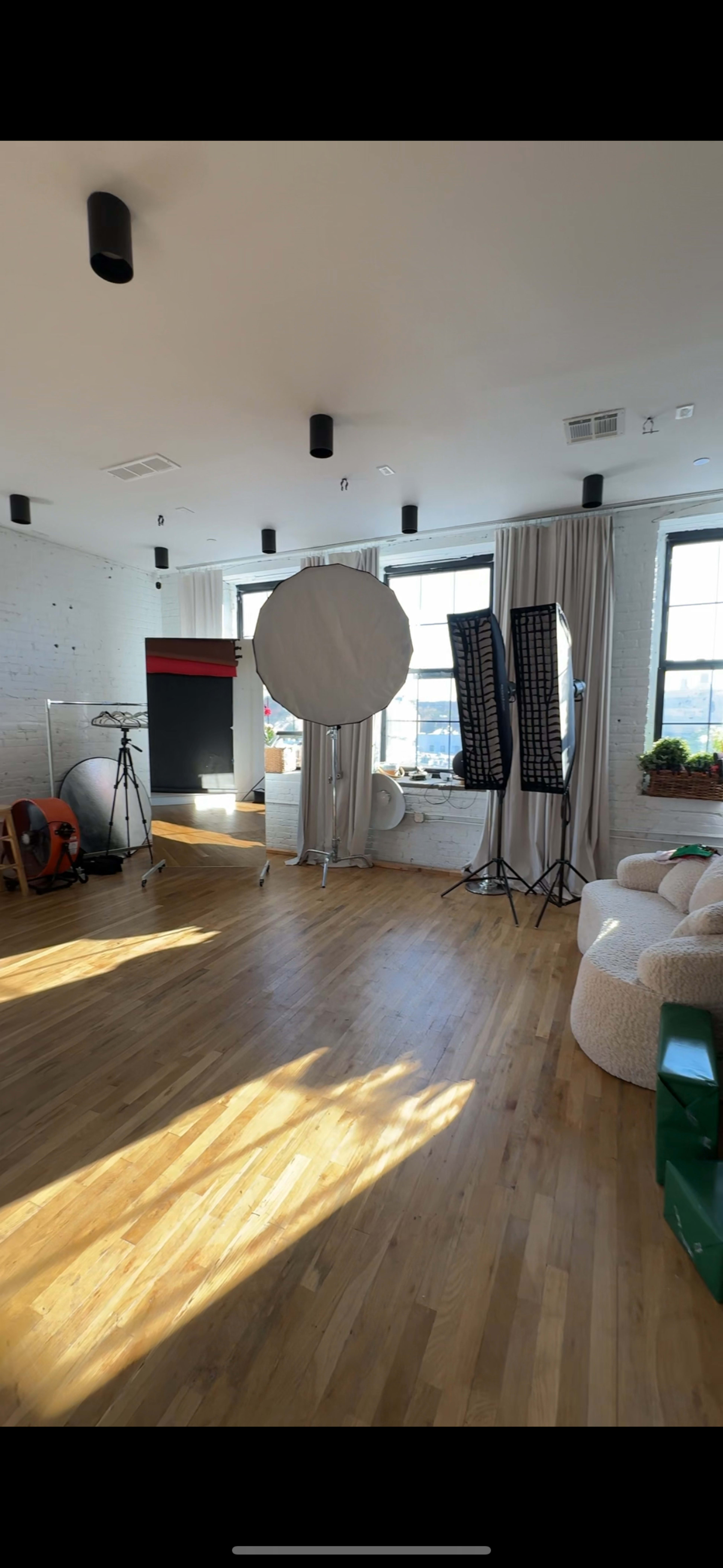 The image shows a well-lit studio space with wooden flooring, featuring several photography equipment items, including light stands, a reflector, and a large couch, with large windows providing natural light.