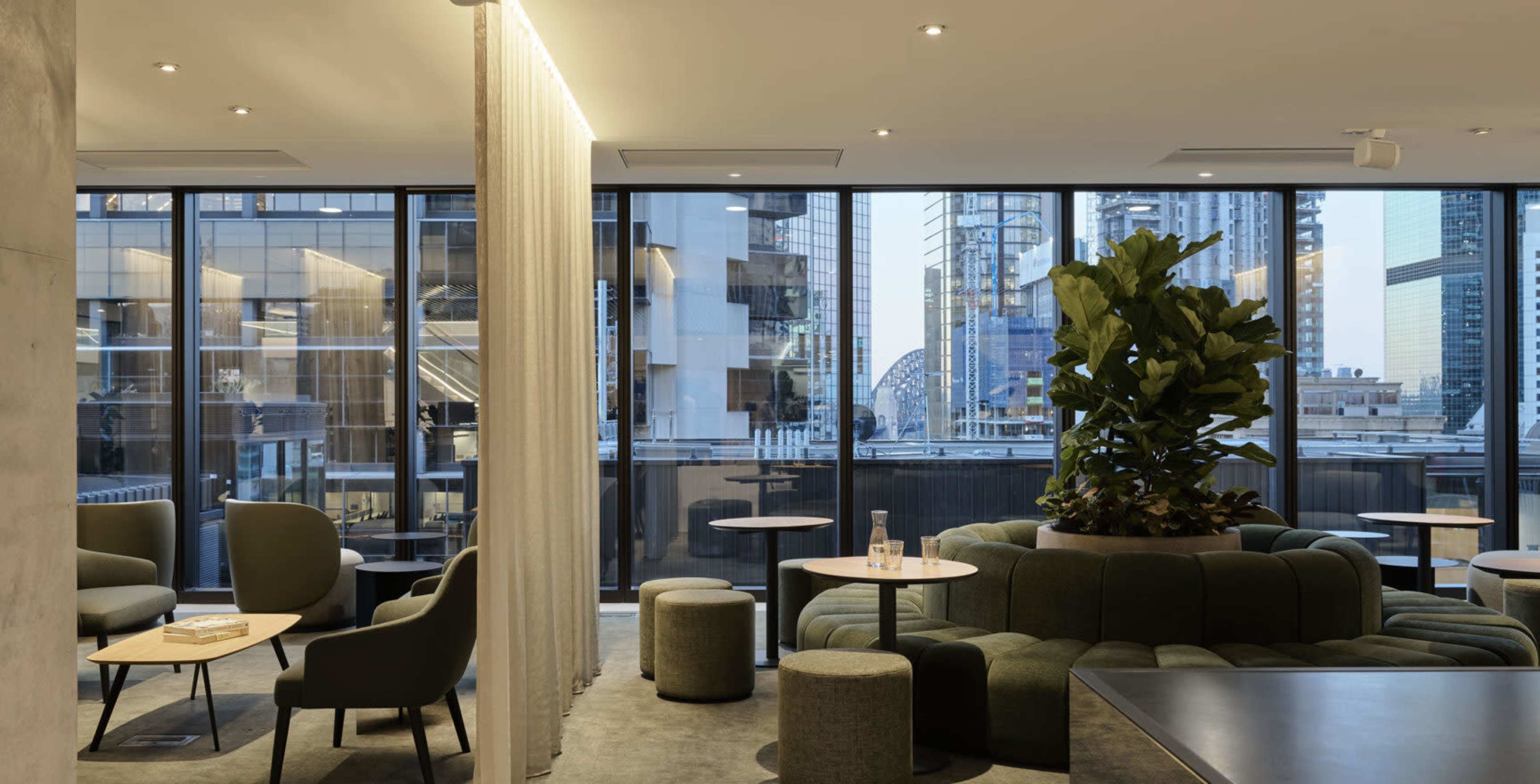 A modern lounge area features green seating, small tables, and large windows with city views.