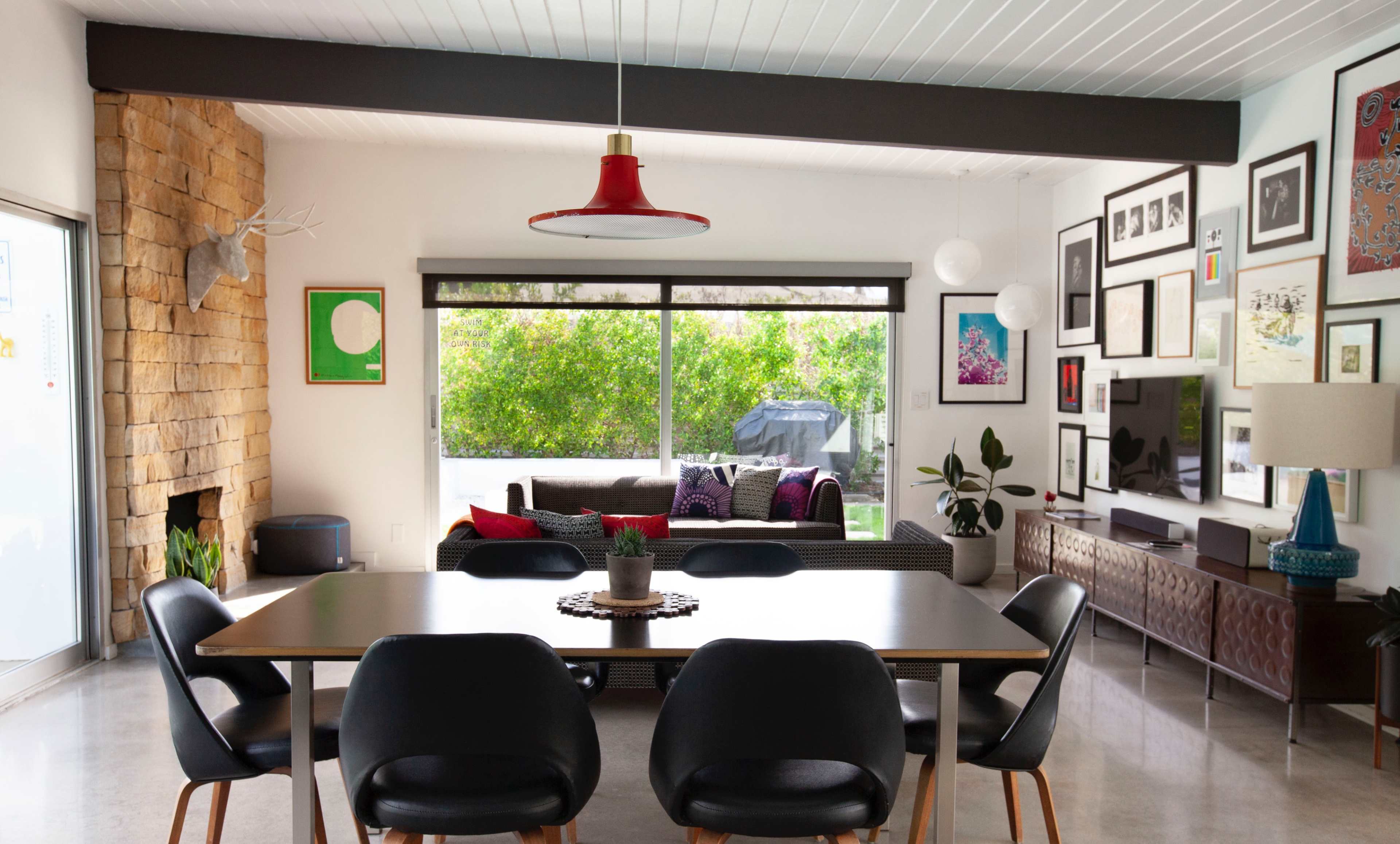 A dining area features a table with black chairs, a stone fireplace, and a large window overlooking greenery.