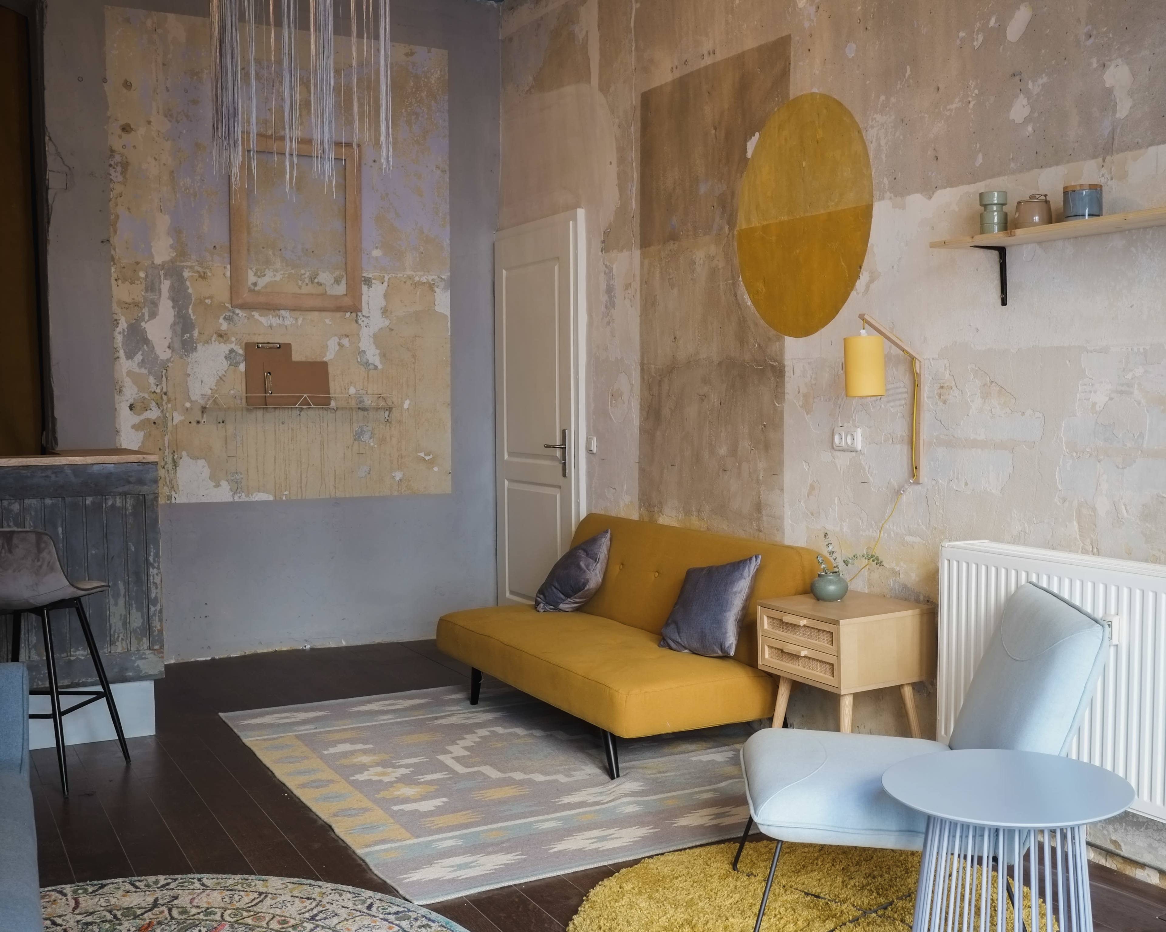 The scene features a sparse living room with a mustard yellow sofa, a light blue chair, and a wooden side table against a wall with peeling paint and geometric patterns.