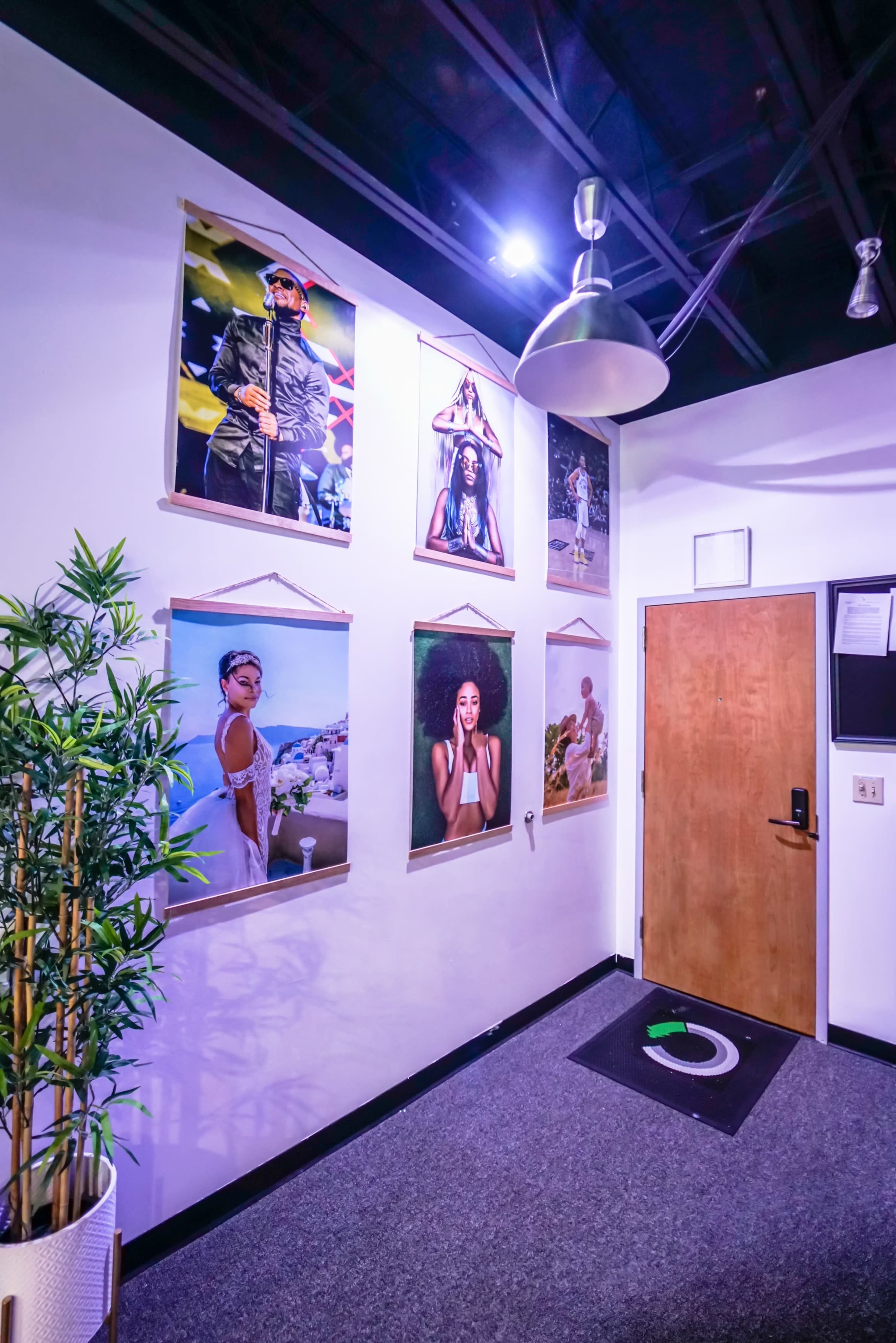 An indoor space featuring a wall adorned with several framed photographs and a wooden door, alongside a potted plant in the corner.