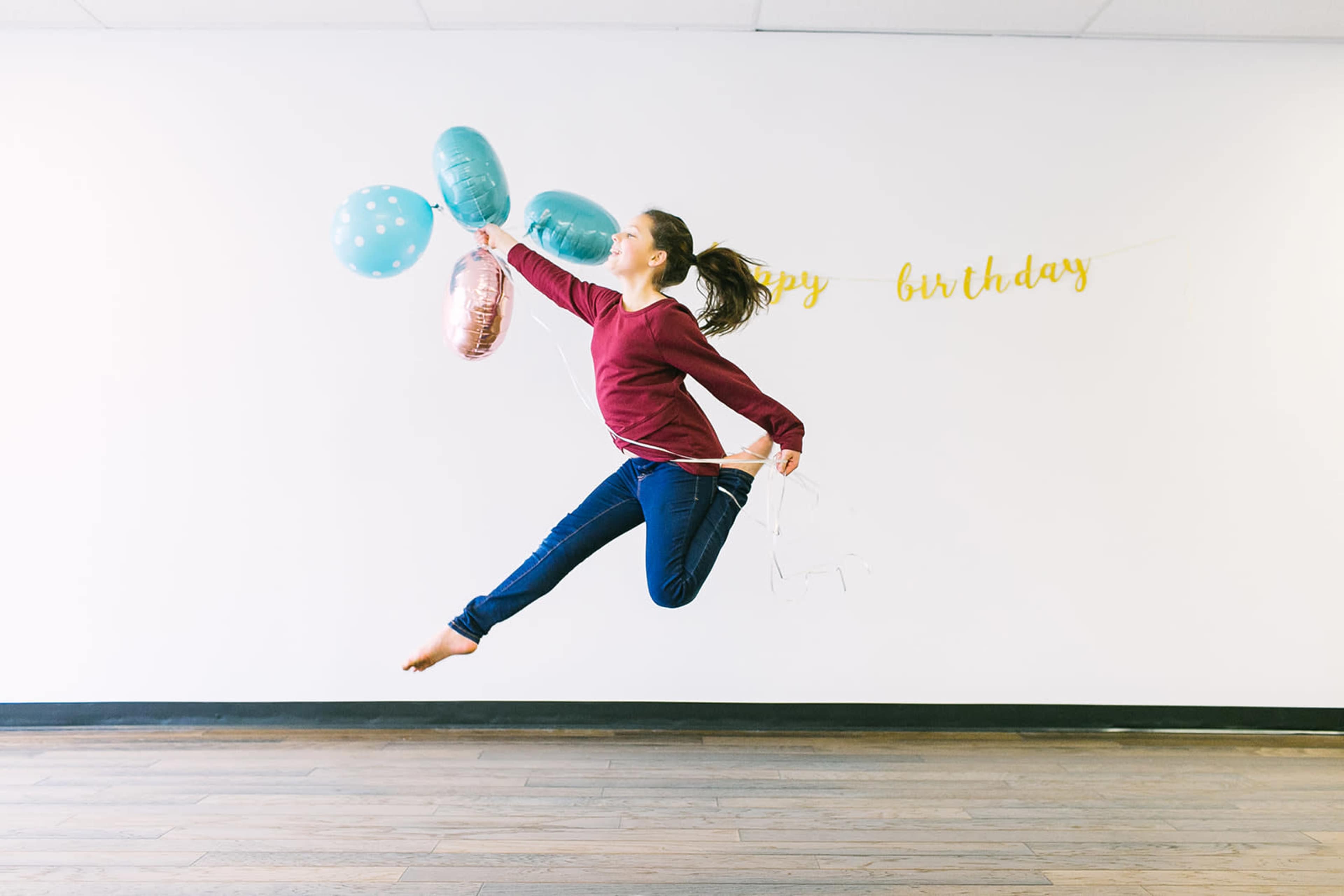 A young girl jumps joyfully while holding a bunch of balloons against a wall decorated with a "Happy Birthday" banner.