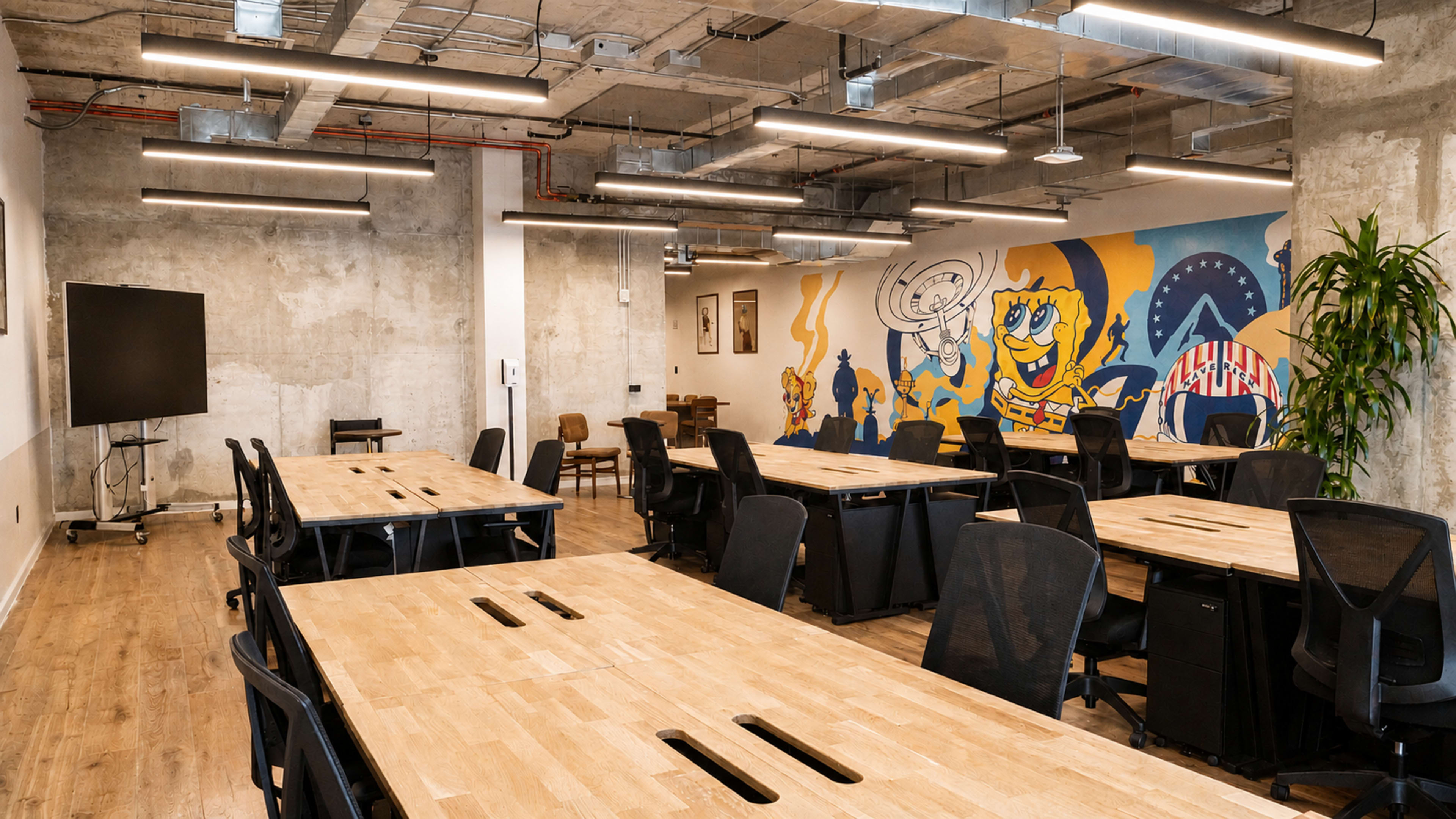 The image shows a modern office space with wooden tables, ergonomic chairs, and a colorful mural featuring cartoon characters on a concrete wall.