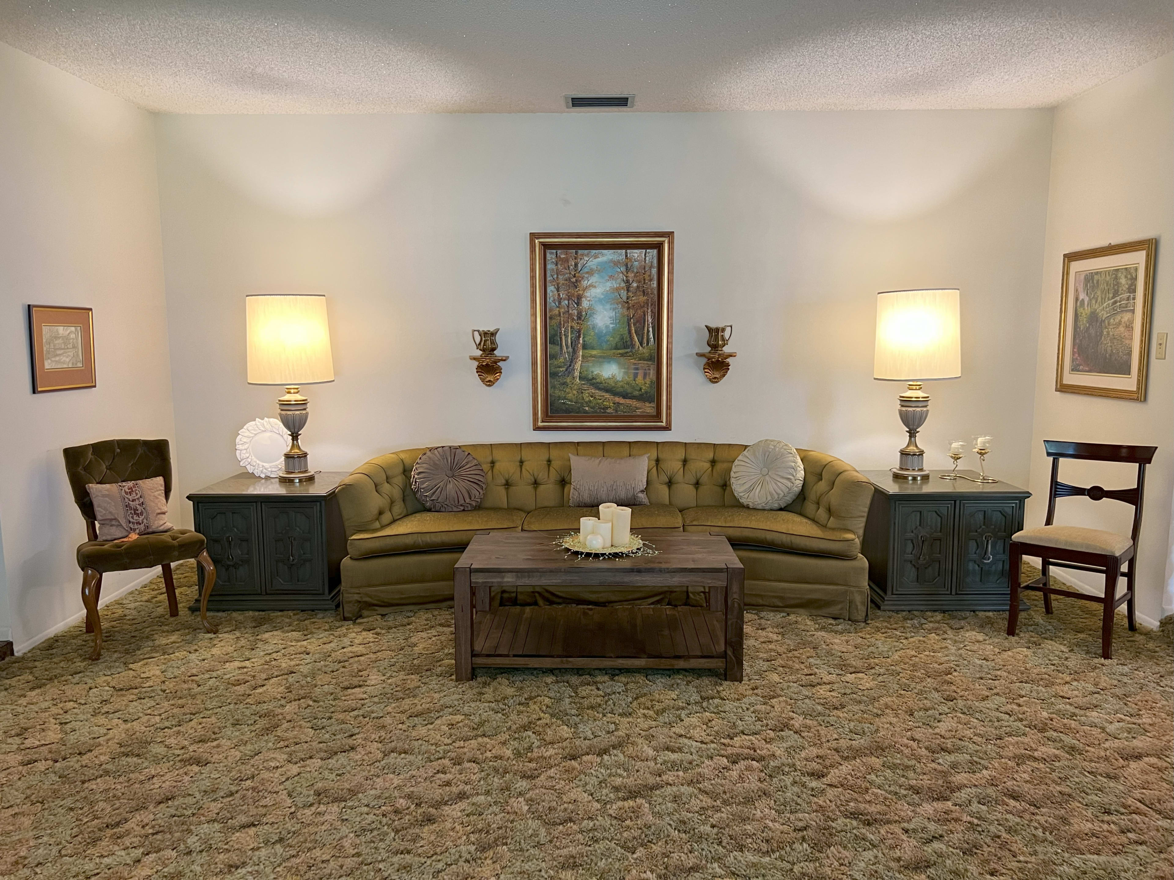 The scene features a living room with an olive green tufted couch, a wooden coffee table in front, and two lamps on either side, accompanied by framed artwork on the walls and patterned carpets.
