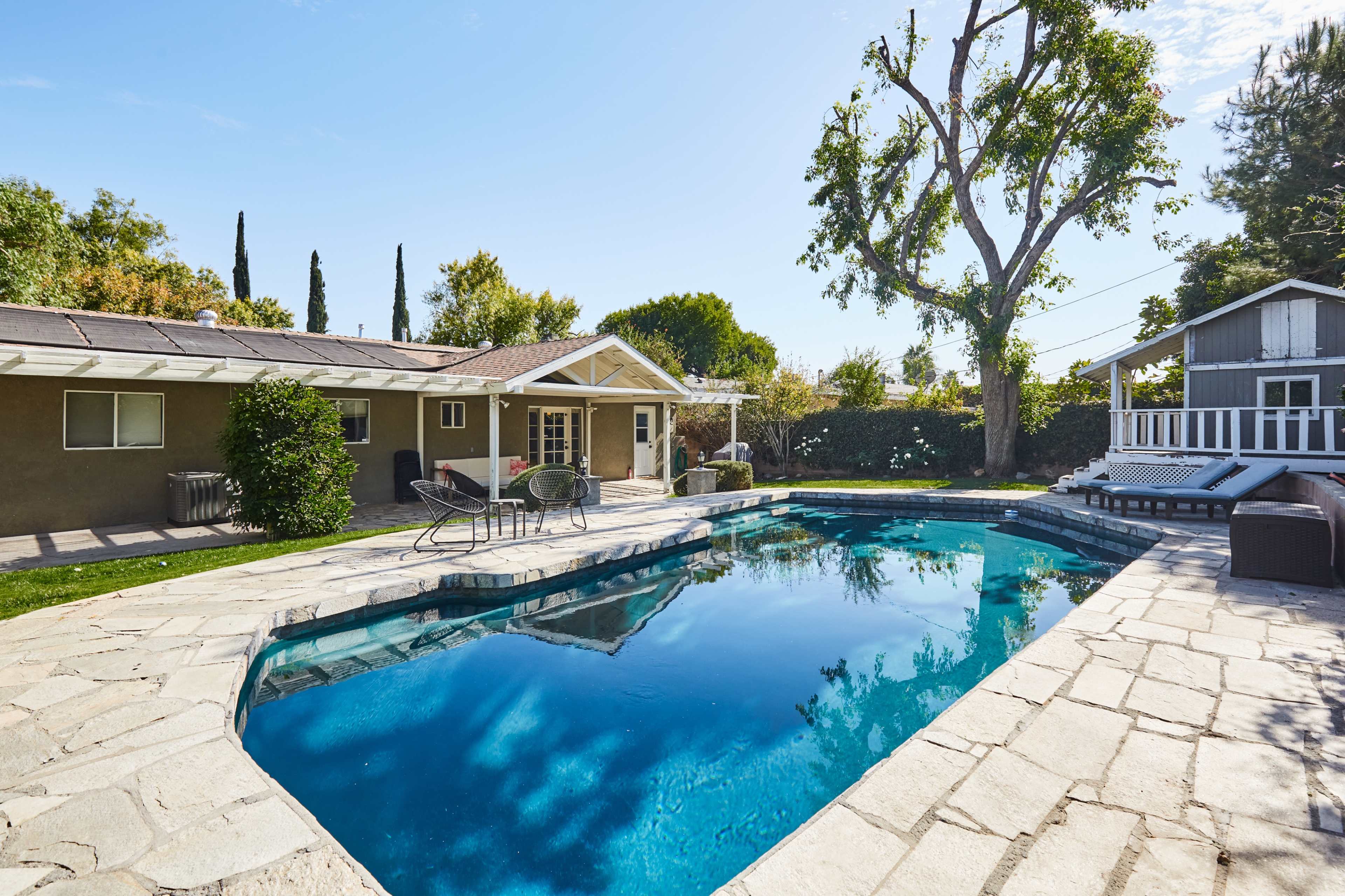A backyard with a rectangular swimming pool surrounded by stone decking, lounge chairs, and a small tree, with a house and a gray shed in the background.