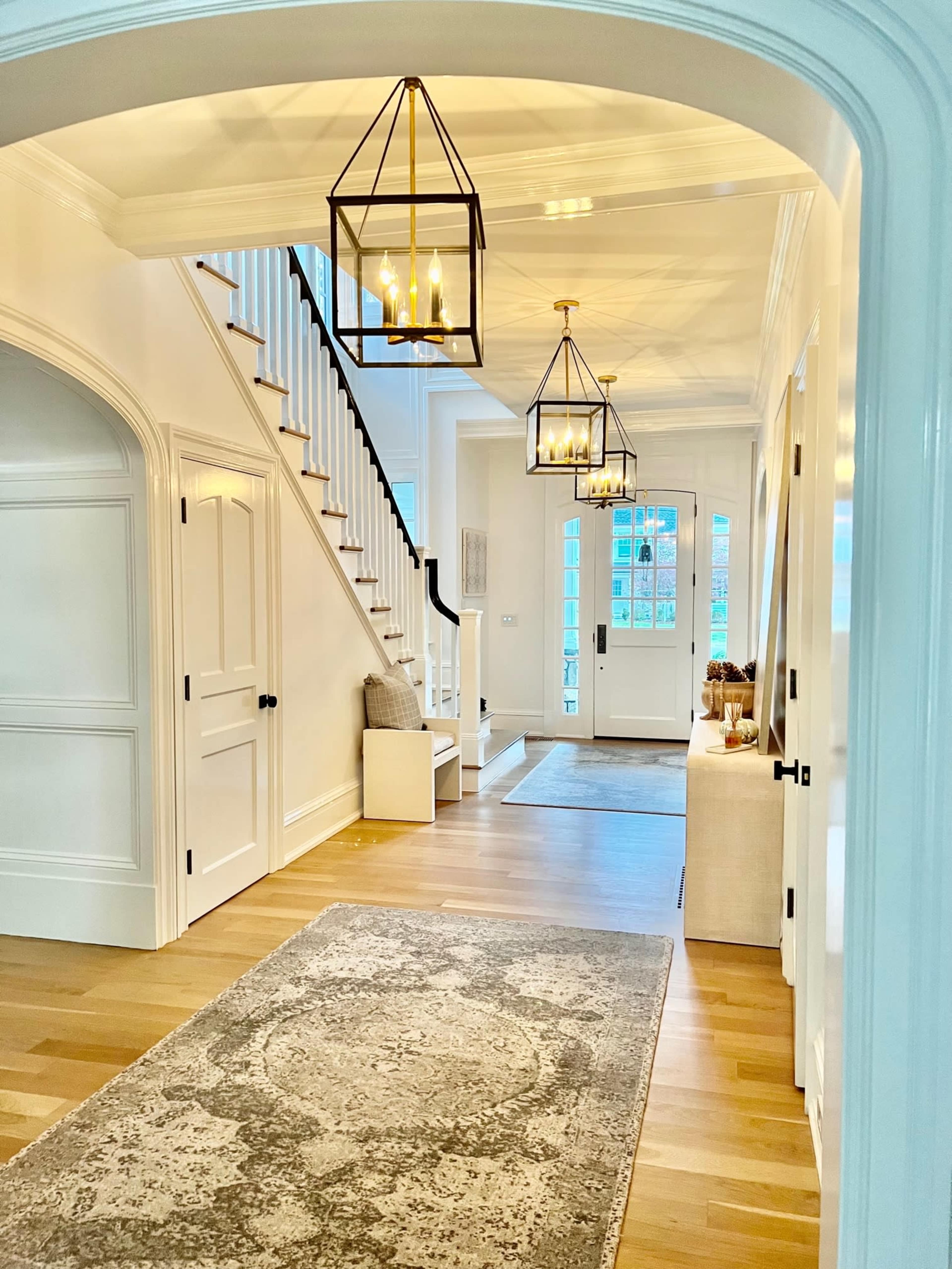 The image depicts a spacious hallway with a staircase, pendant lighting, and a large rug on a wooden floor.