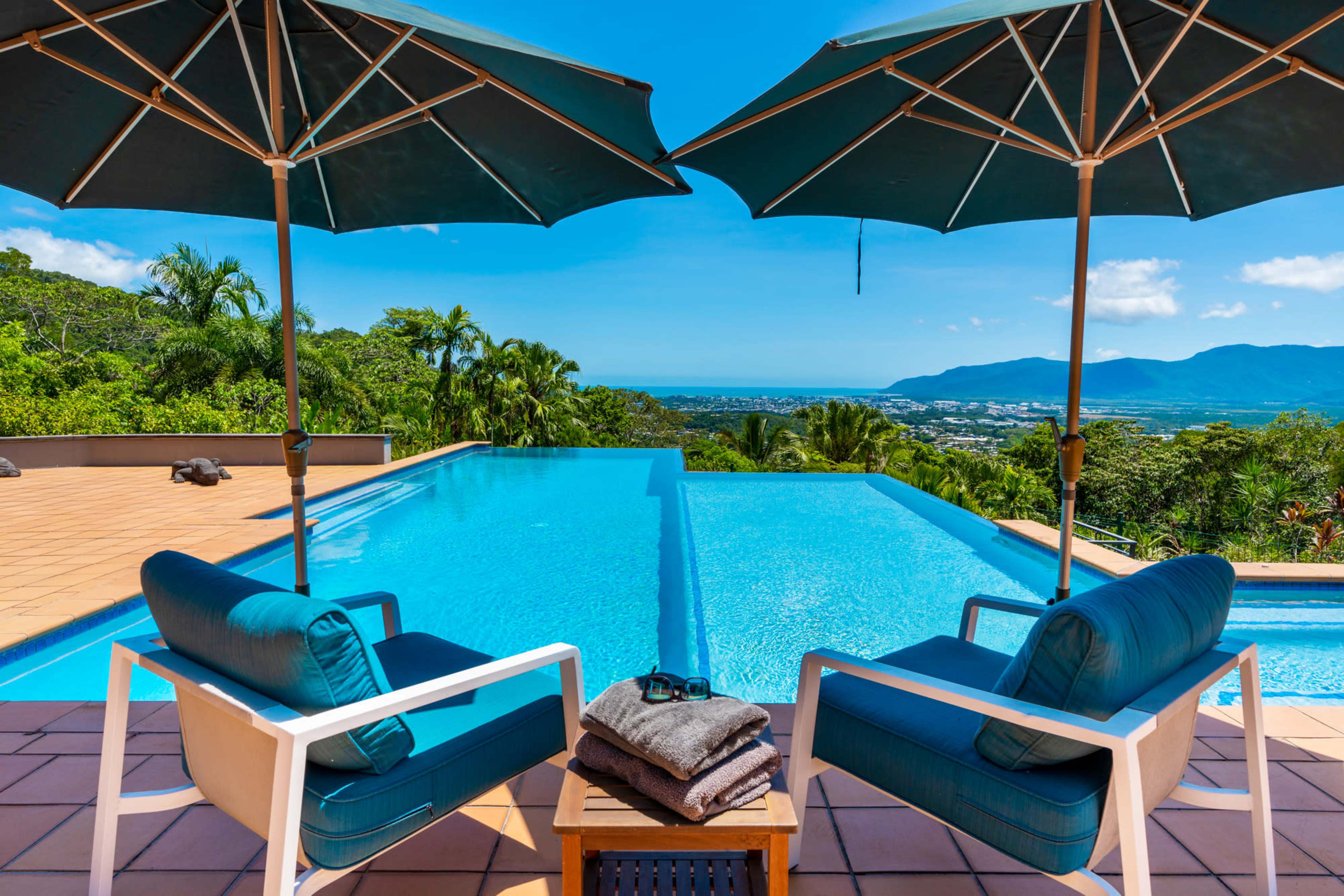 Two chairs with blue cushions and a wooden table sit beside an infinity pool, overlooking a lush landscape and blue skies with distant mountains.