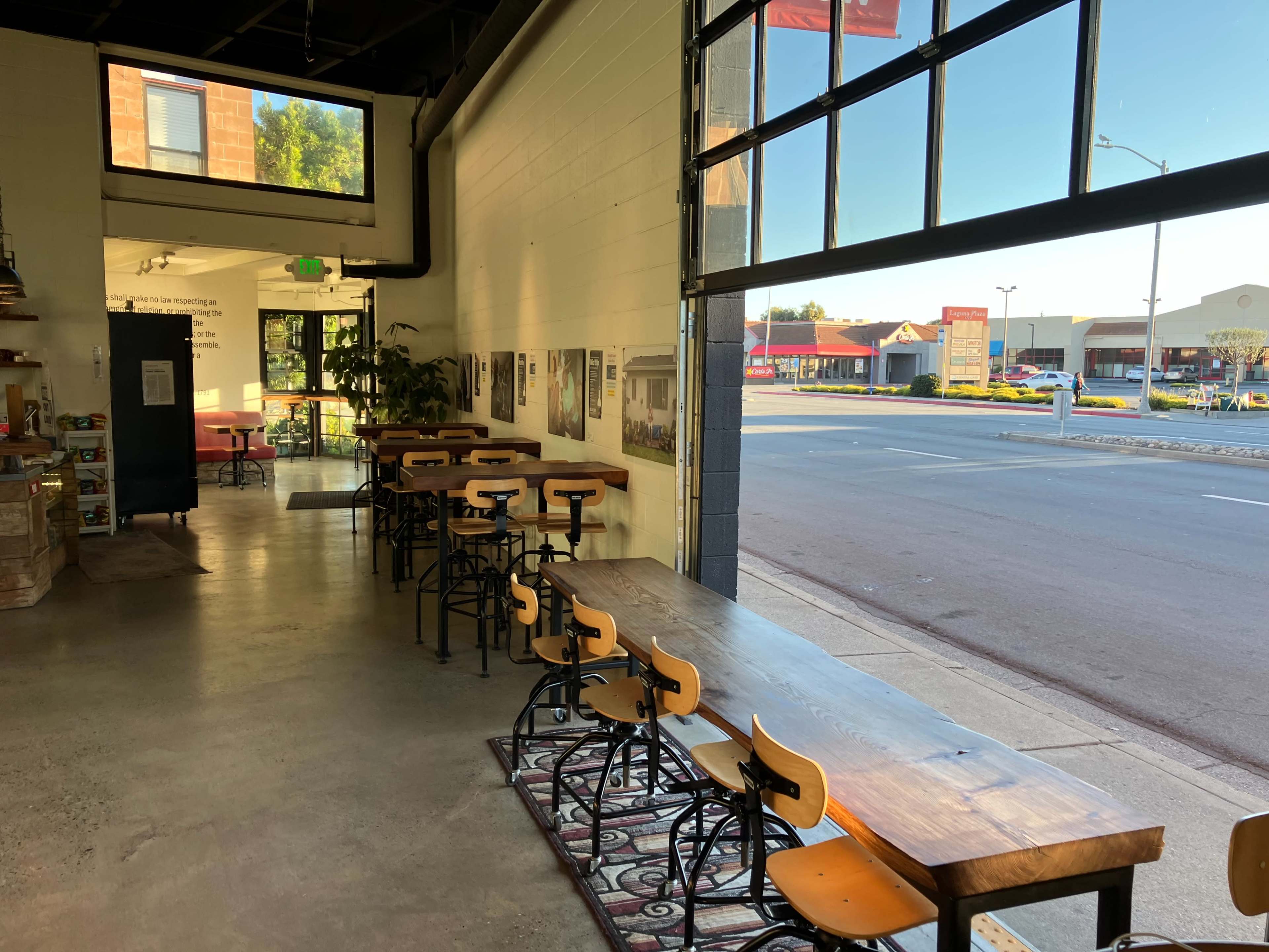 A coffee shop interior featuring wooden tables and chairs along one side, with large windows facing the street.