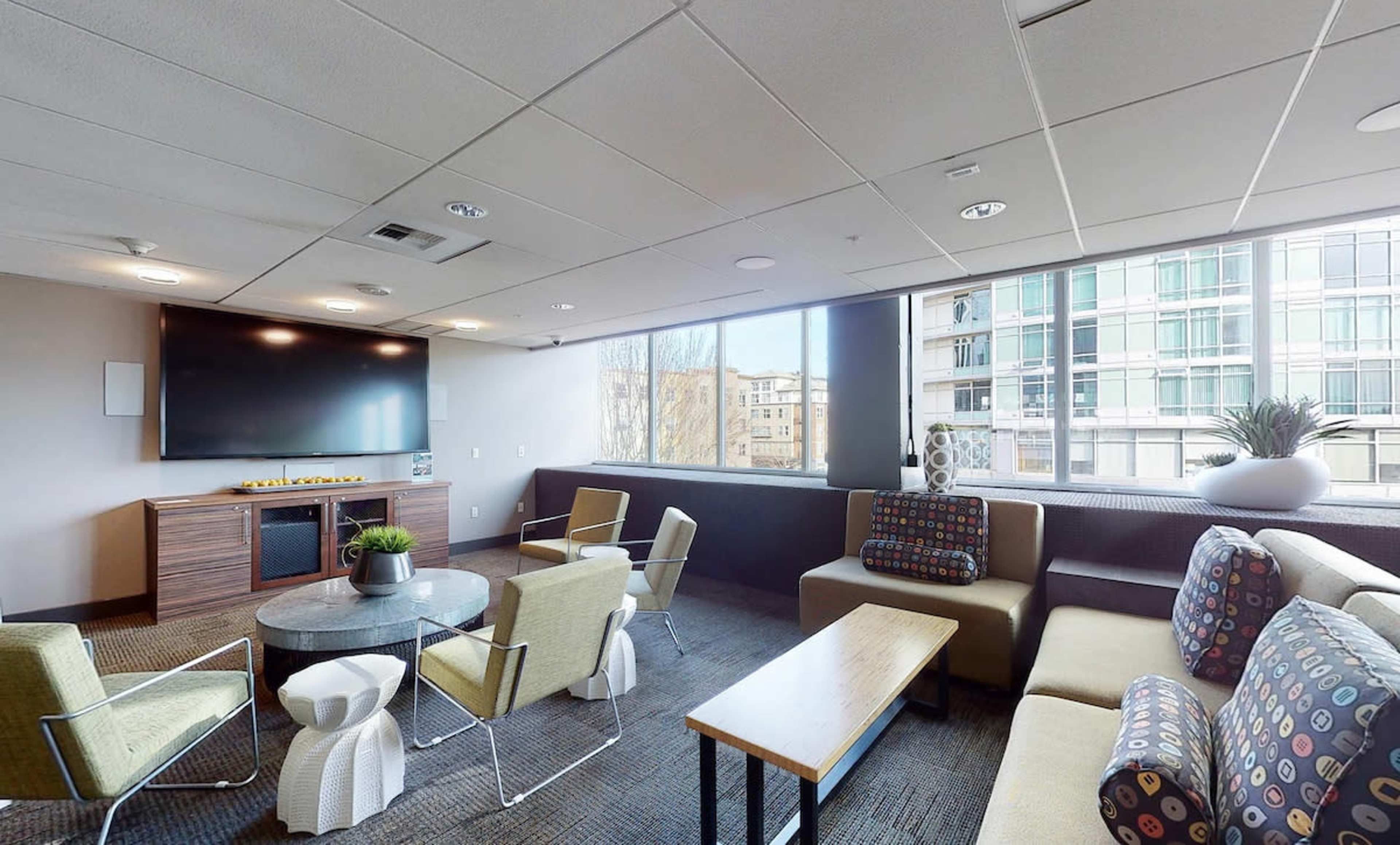 The image shows a modern lounge area with comfortable seating, a television, and large windows allowing natural light to enter.