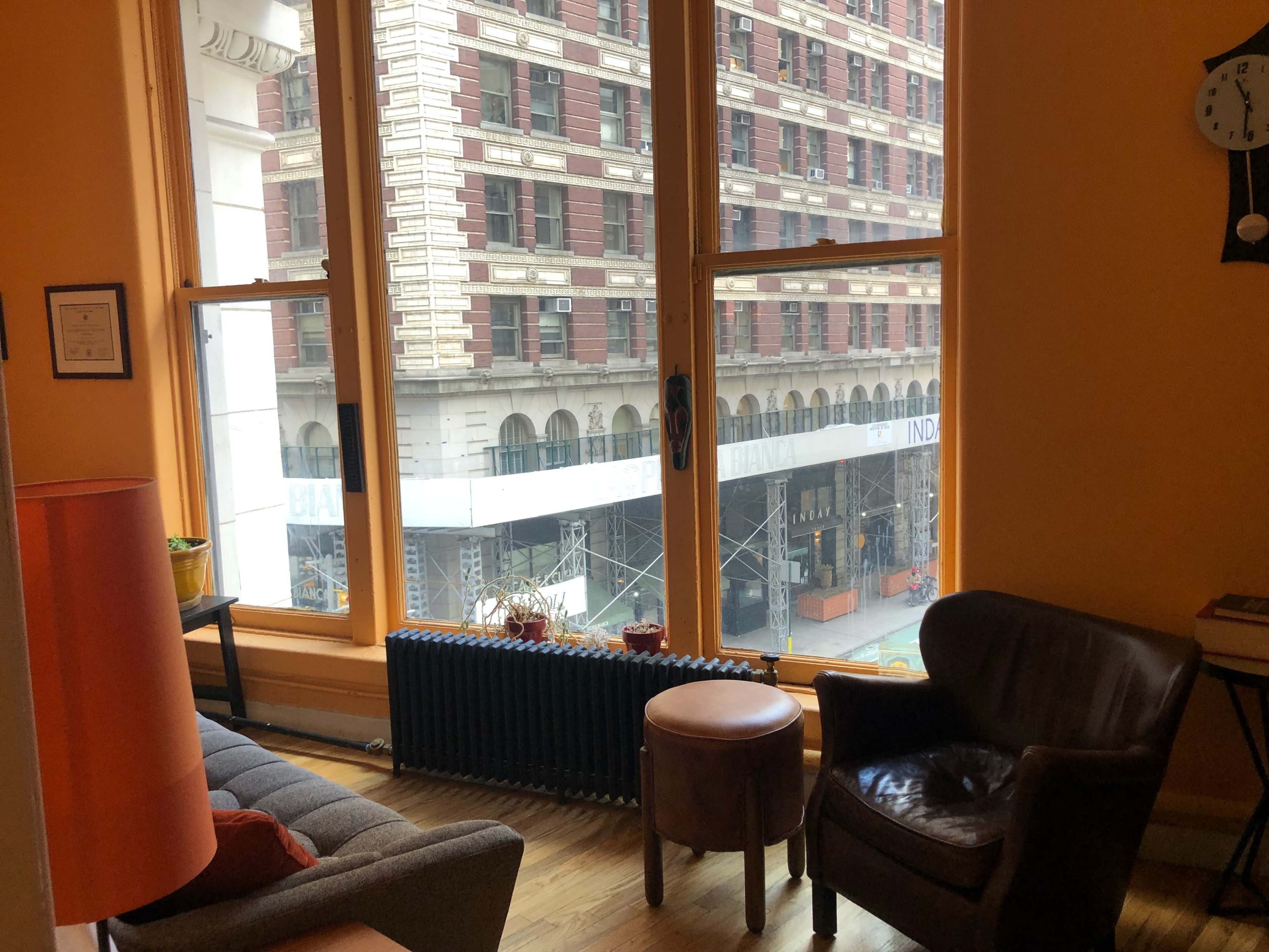 A cozy room with a gray sofa, a wooden table, and large windows displaying a view of a building under renovation.