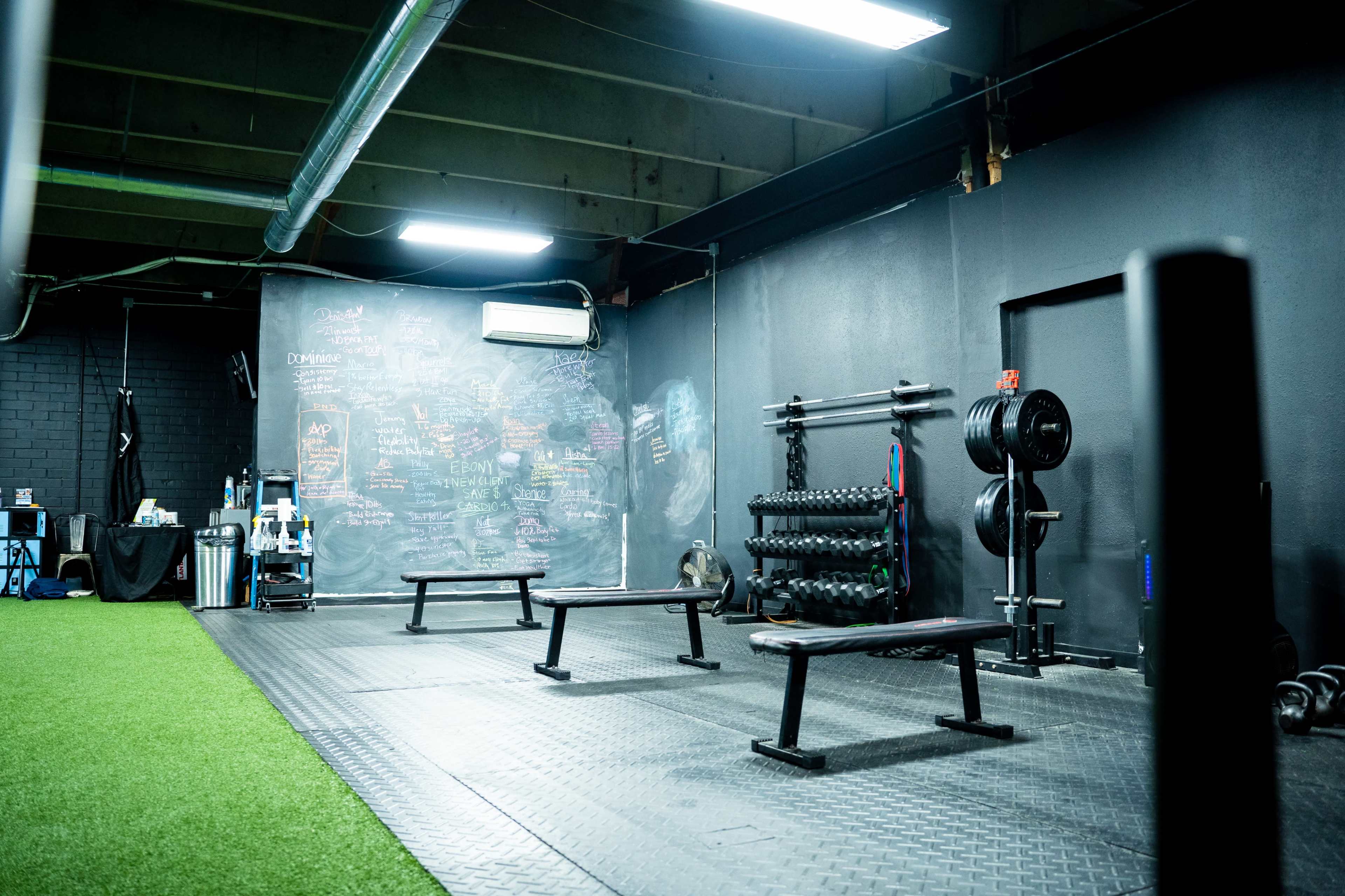 A gym space with several workout benches, dumbbells on a rack, and a chalkboard wall filled with notes, all on a rubber floor.