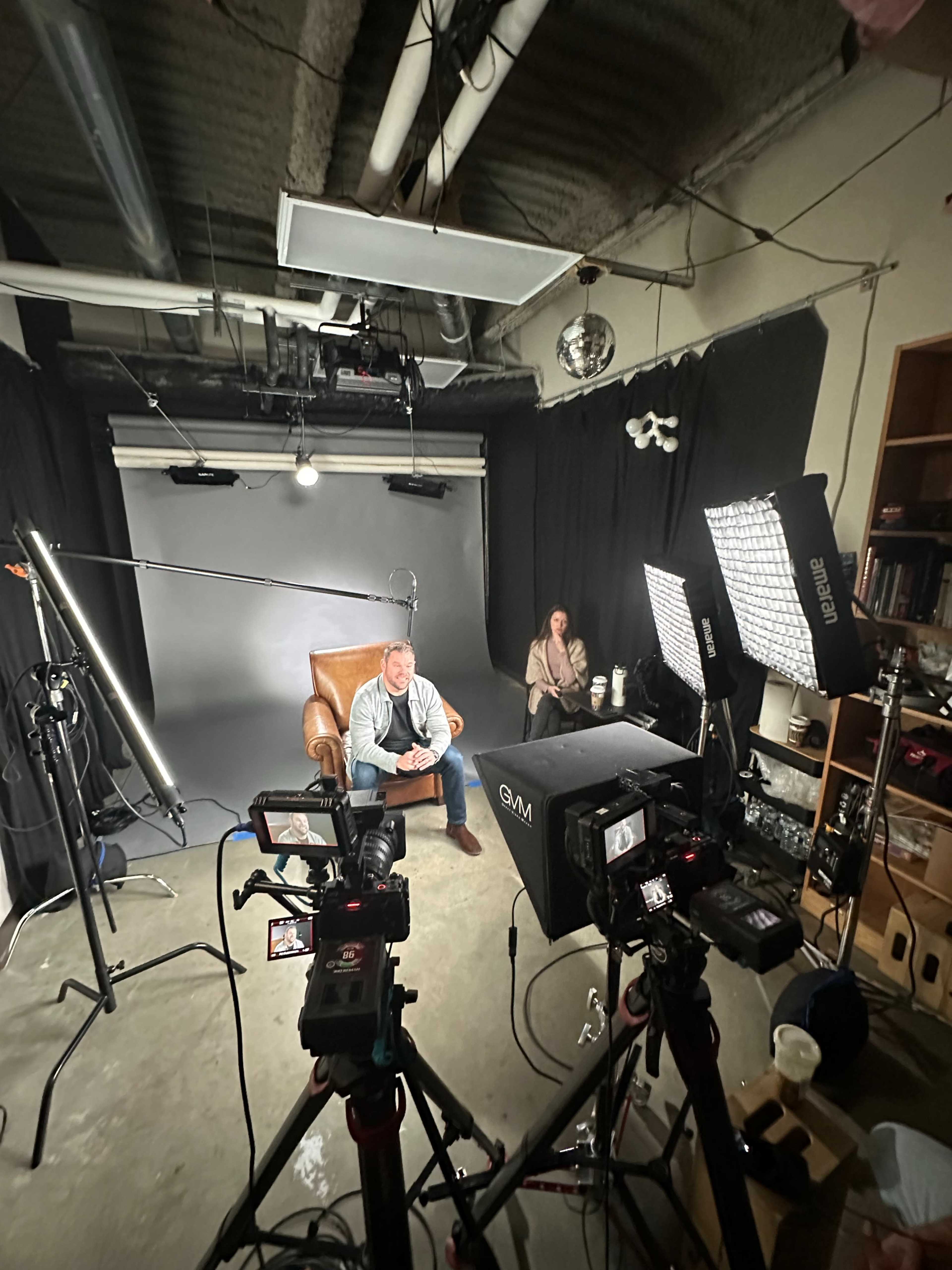 A man sits in a chair in a studio setting surrounded by cameras and lighting equipment, while a woman is positioned nearby.