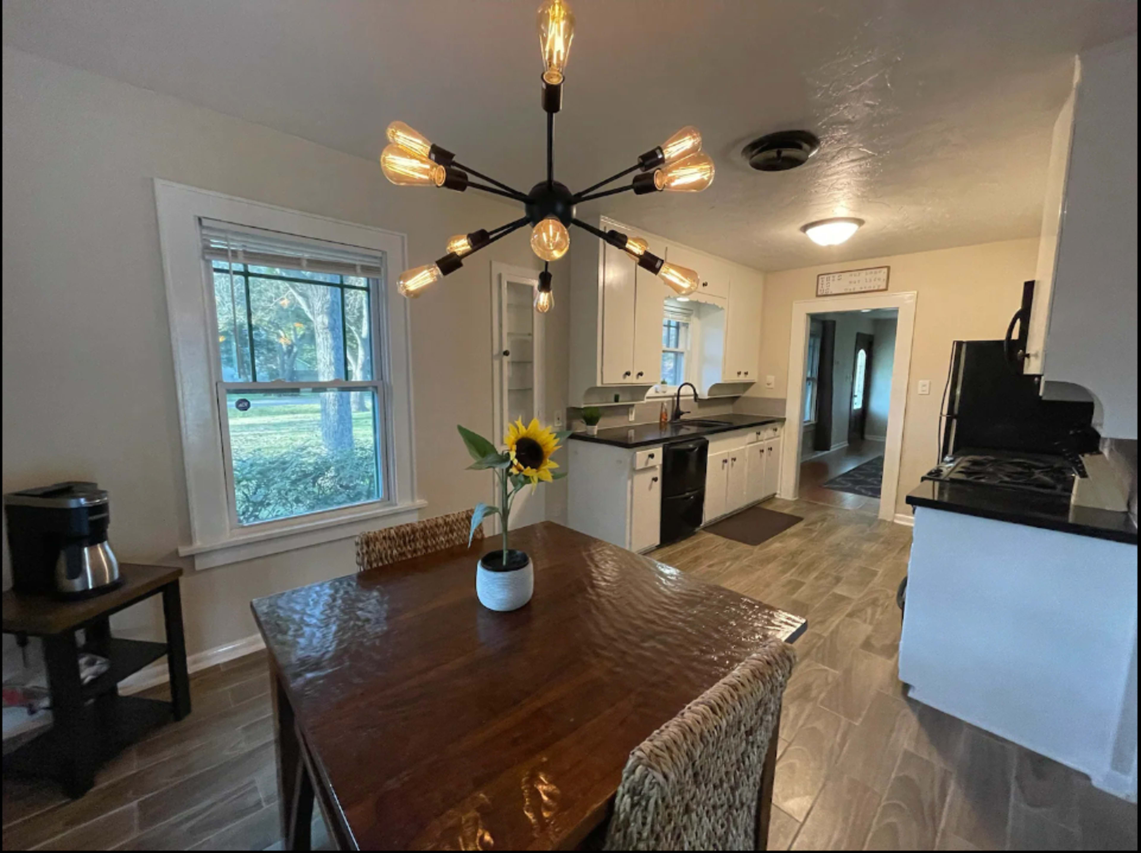 A kitchen features a wooden dining table with a sunflower centerpiece, a black countertop, and a modern light fixture overhead.