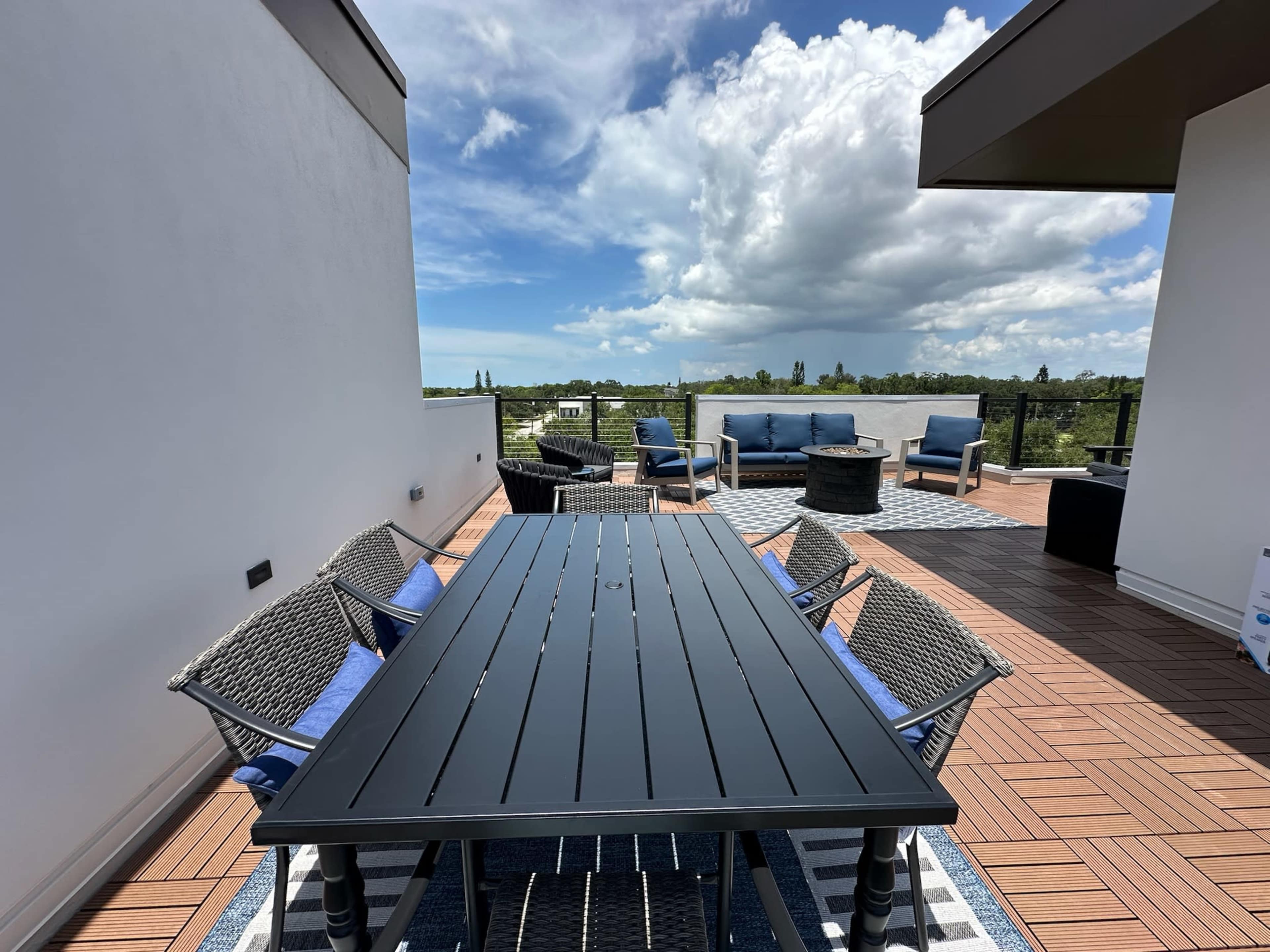A rooftop patio features a long black dining table surrounded by wicker chairs and a separate seating area with blue cushions, under a partly cloudy sky.