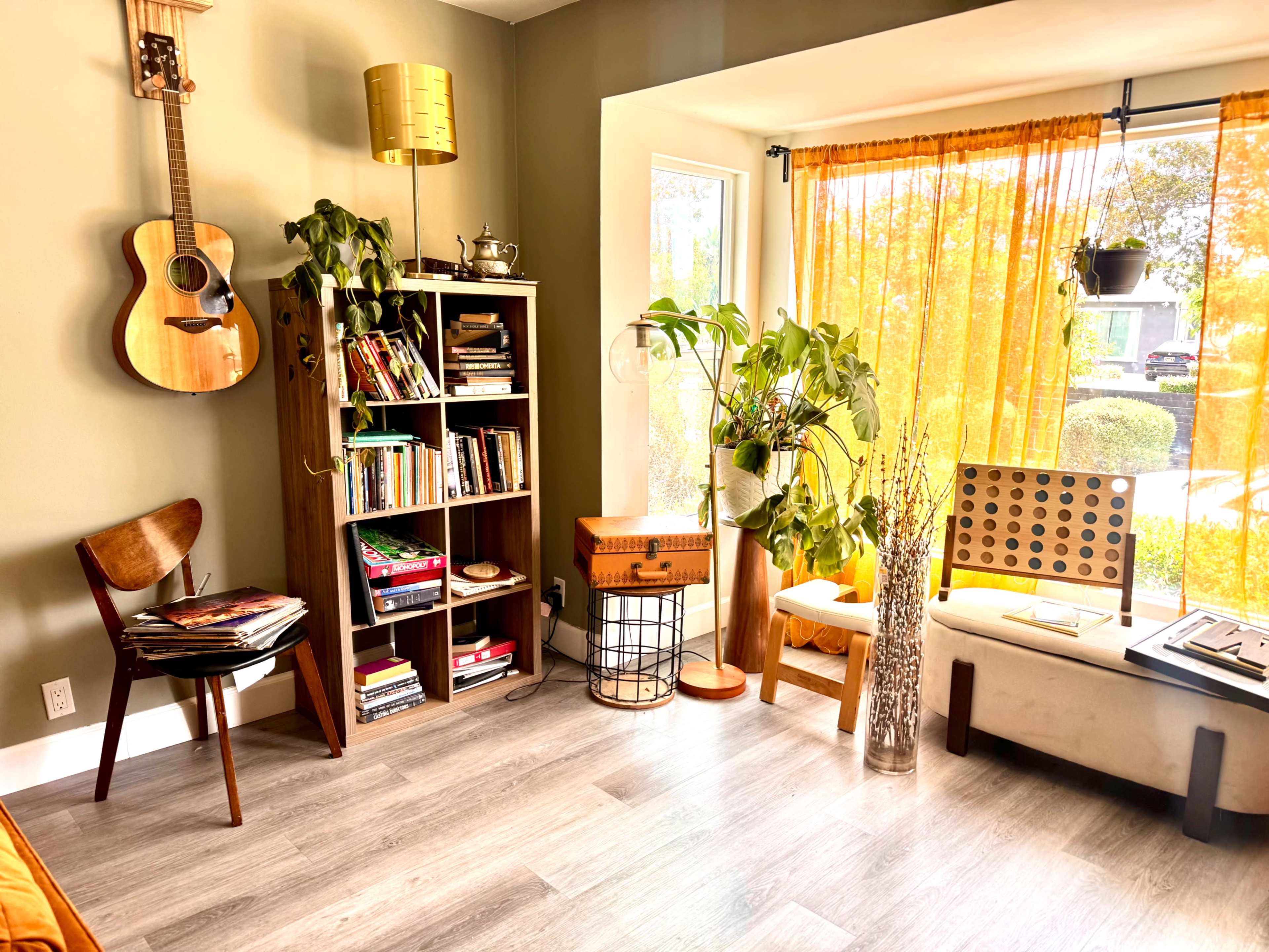 A cozy living room features a guitar on the wall, a bookshelf filled with books, various plants, and natural light streaming through large windows.