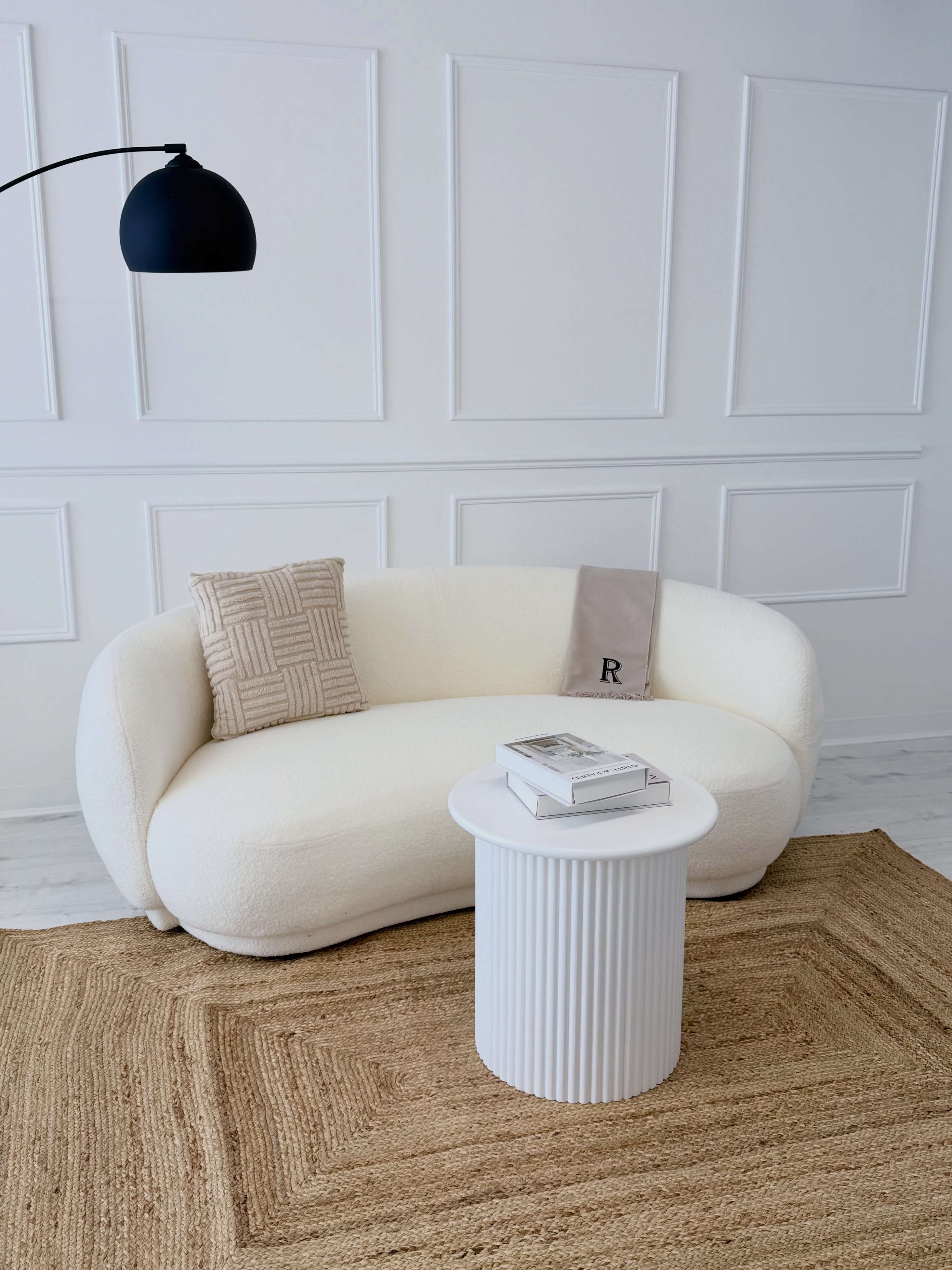 A plush, white curved sofa is positioned next to a round white table on a textured beige rug in a minimalist room with paneled walls.
