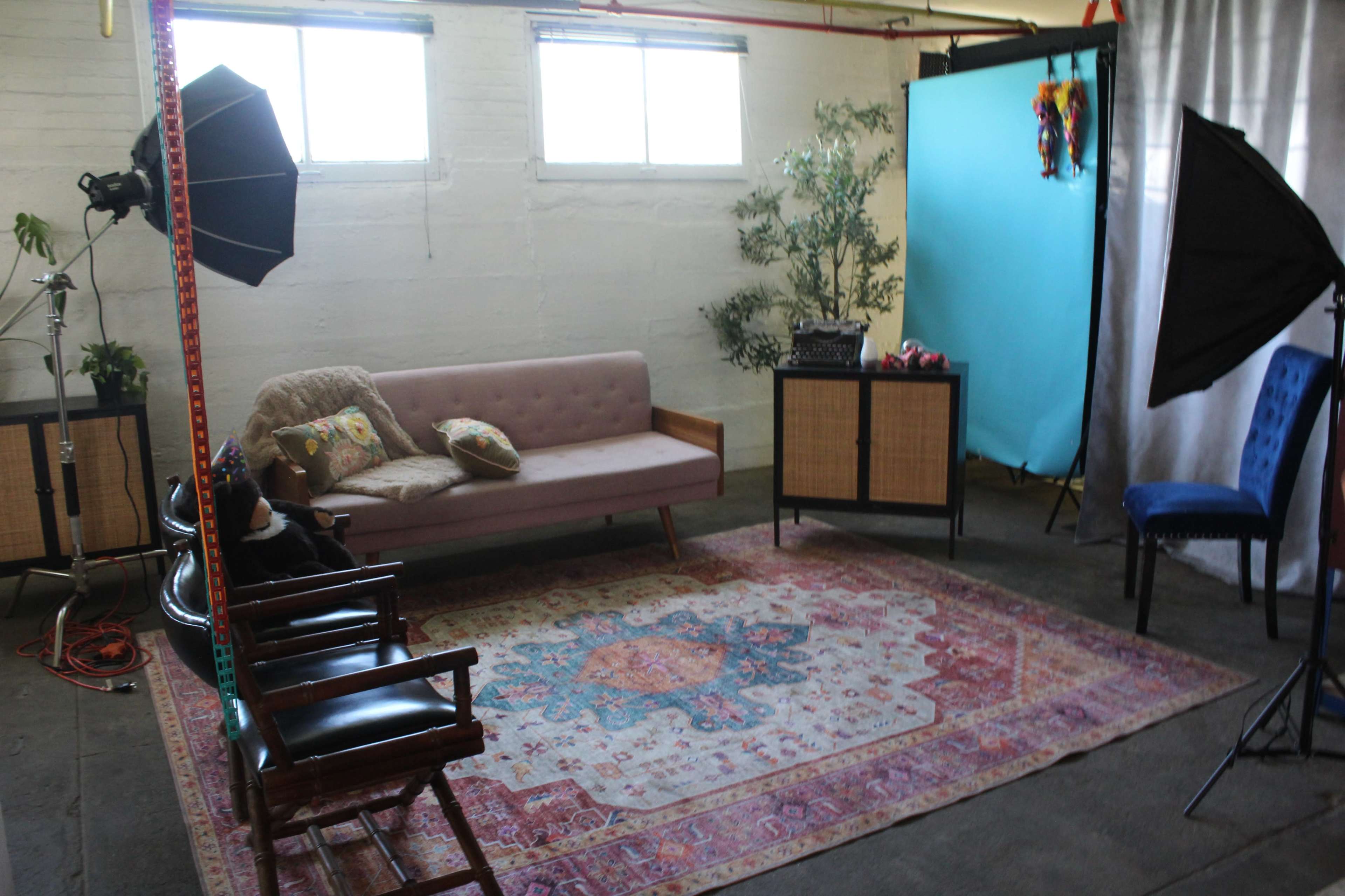 The image shows a photography studio with a pink sofa, a patterned rug, two softbox lights, a blue backdrop, and various props arranged in the space.