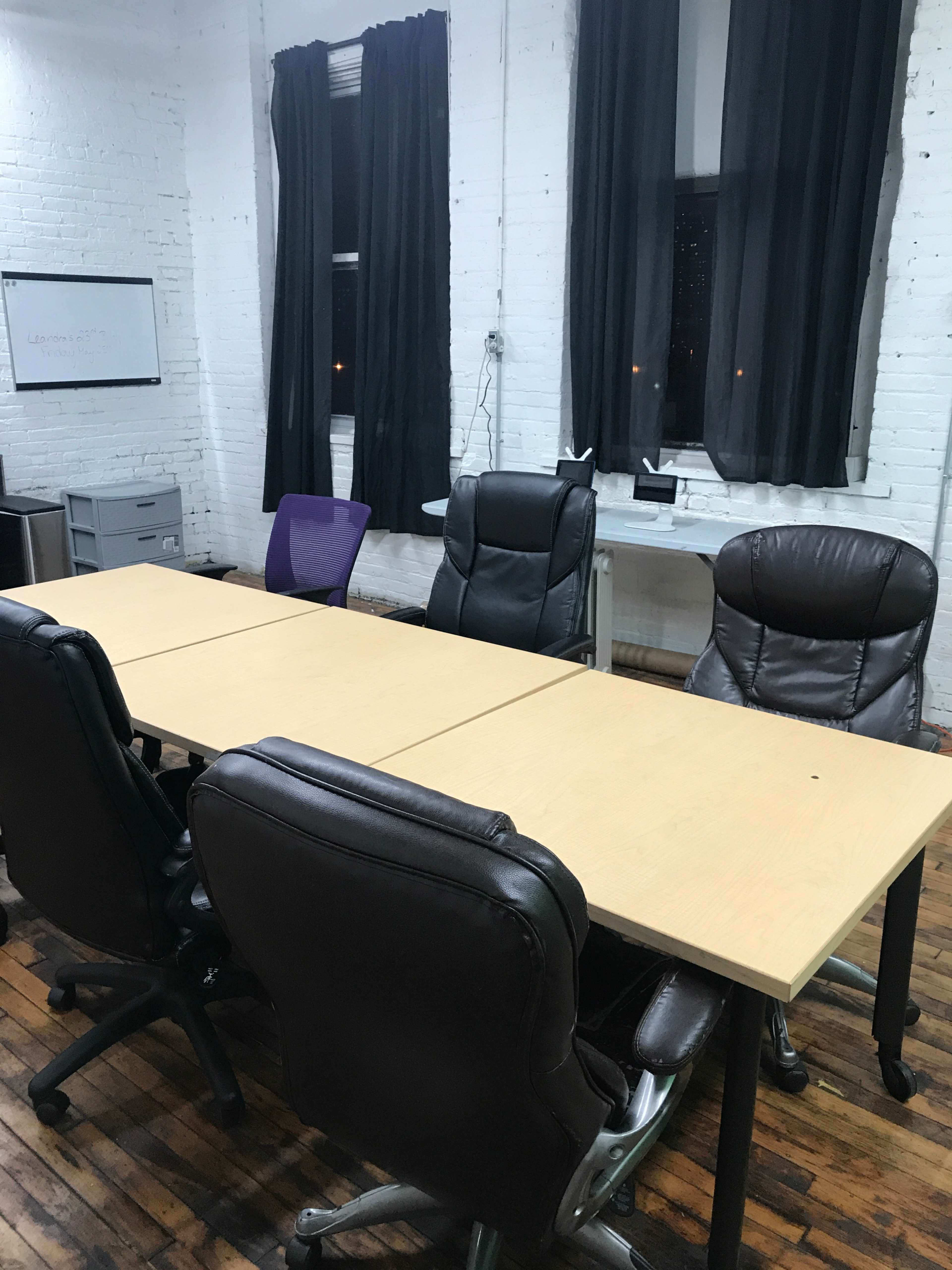 A long wooden conference table with five chairs is set up in a room with white brick walls and large black curtains covering the windows.