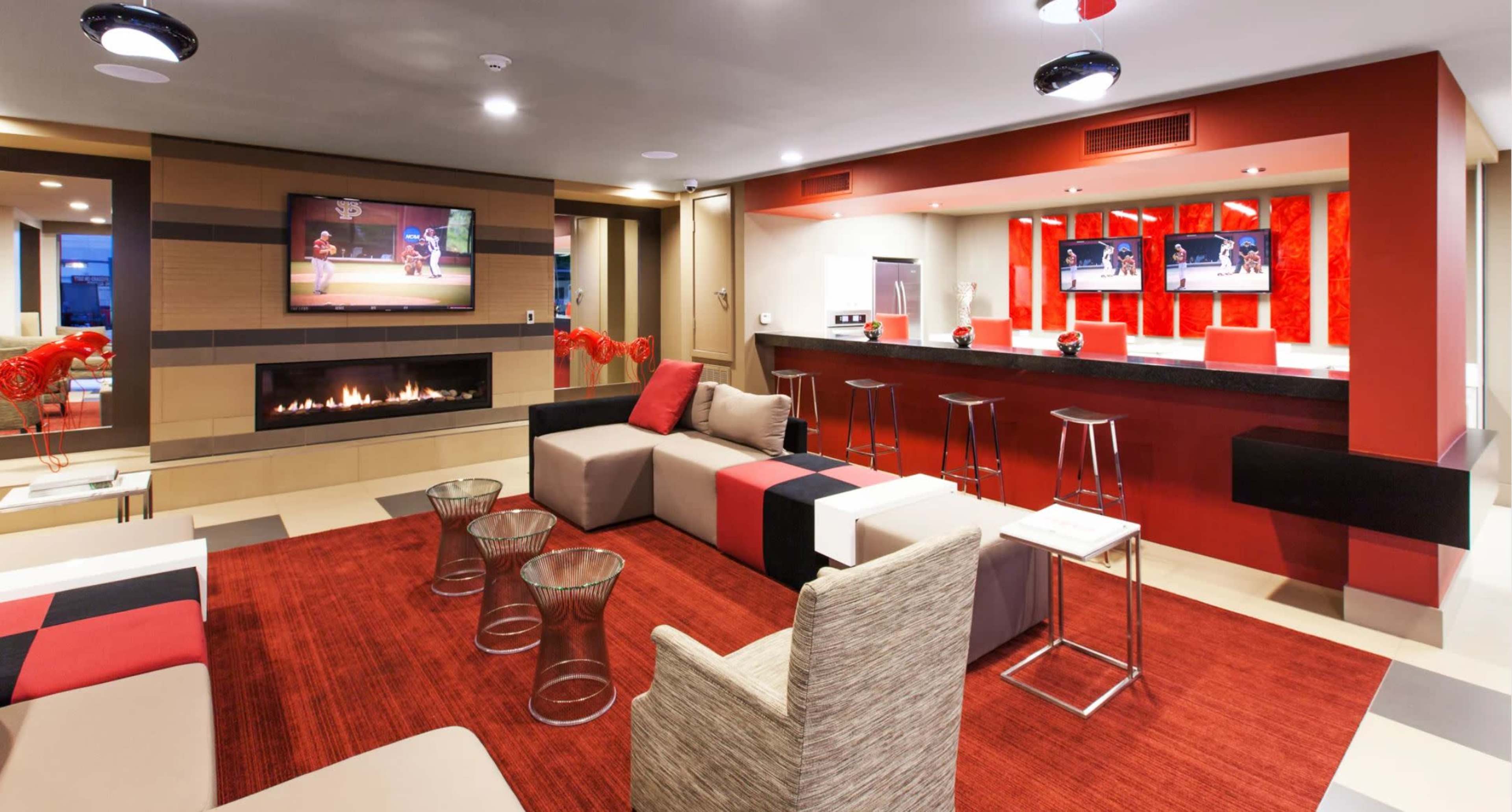 The image shows a modern lounge area featuring a fireplace, a large television, a bar with high stools, and various seating arrangements on a red and neutral-toned rug.