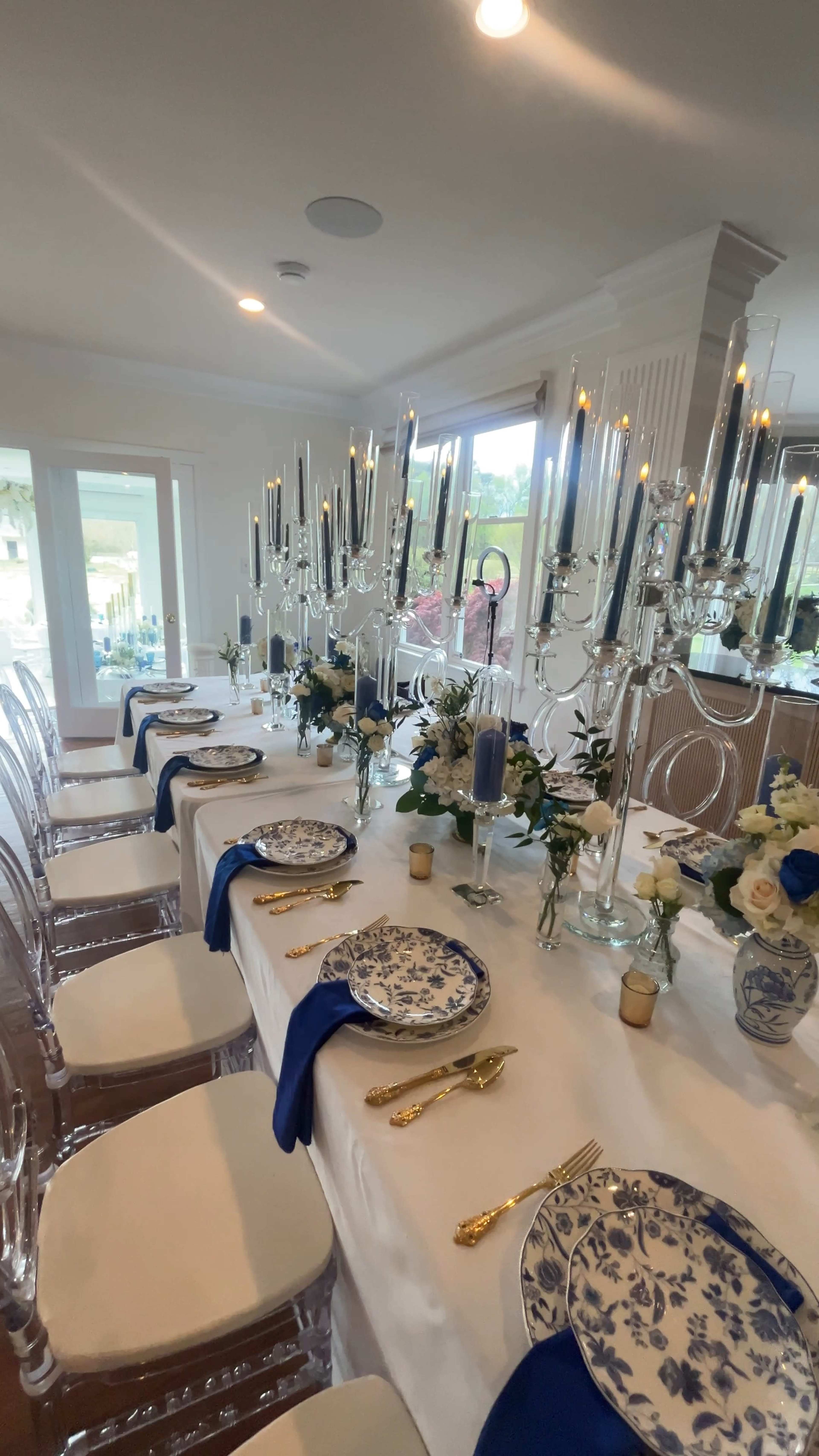 A long dining table is elegantly set with blue and white dishes, gold utensils, and tall candelabras surrounded by floral arrangements.