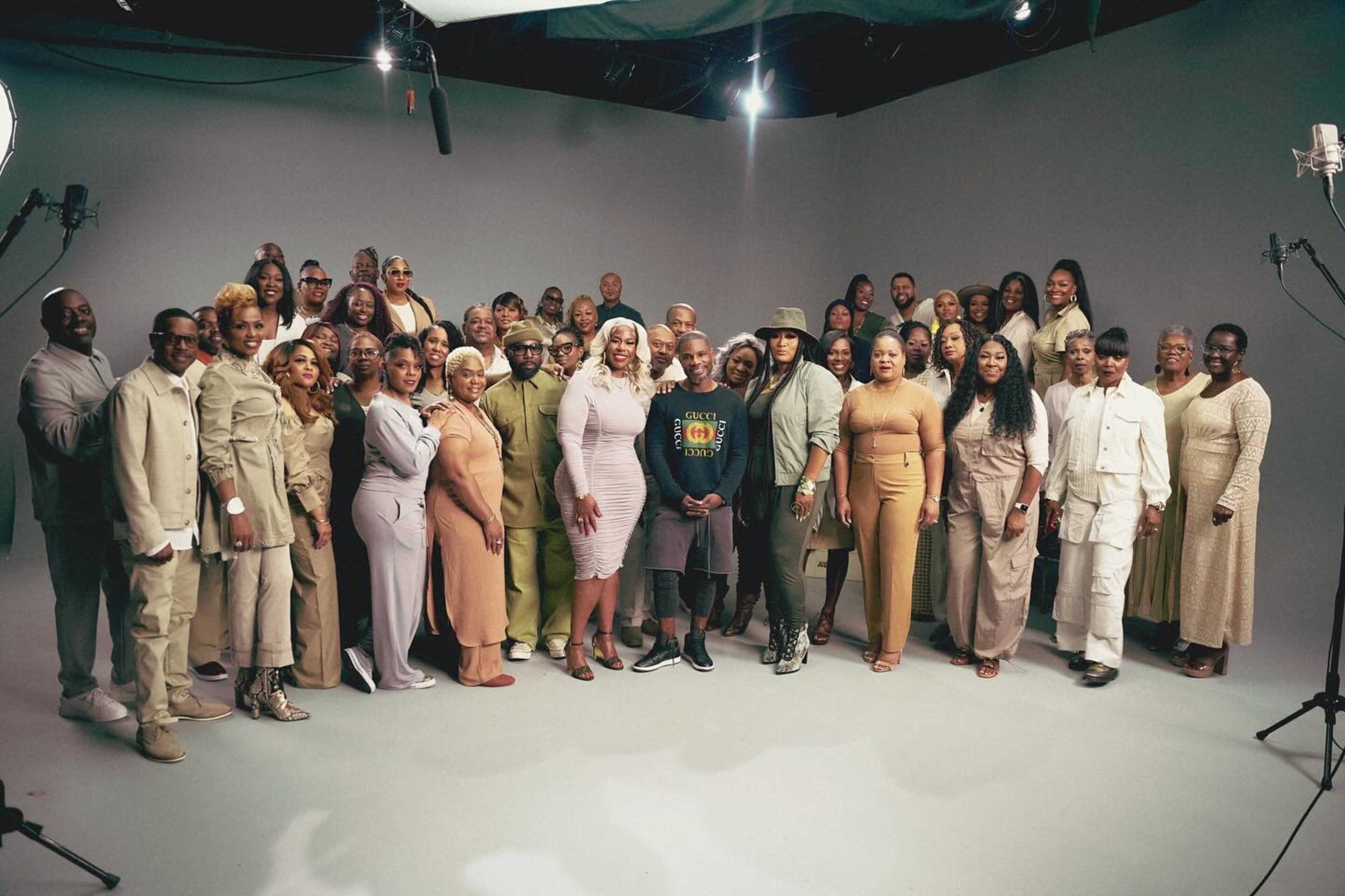 A large group of diverse individuals is gathered together in a studio setting, standing in front of a plain backdrop.