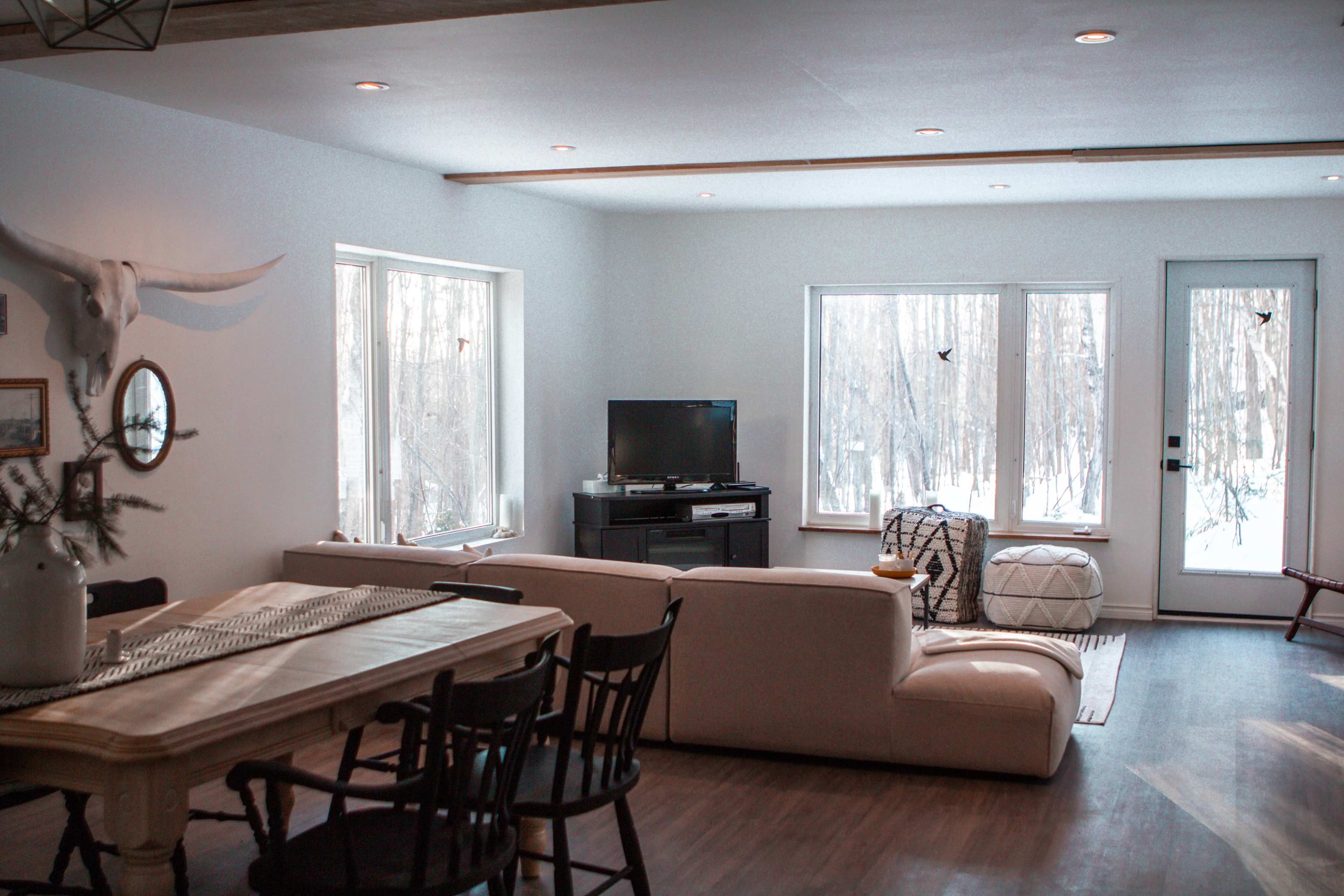 A modern living room with a white couch, a wooden dining table, and large windows overlooking a snowy landscape.