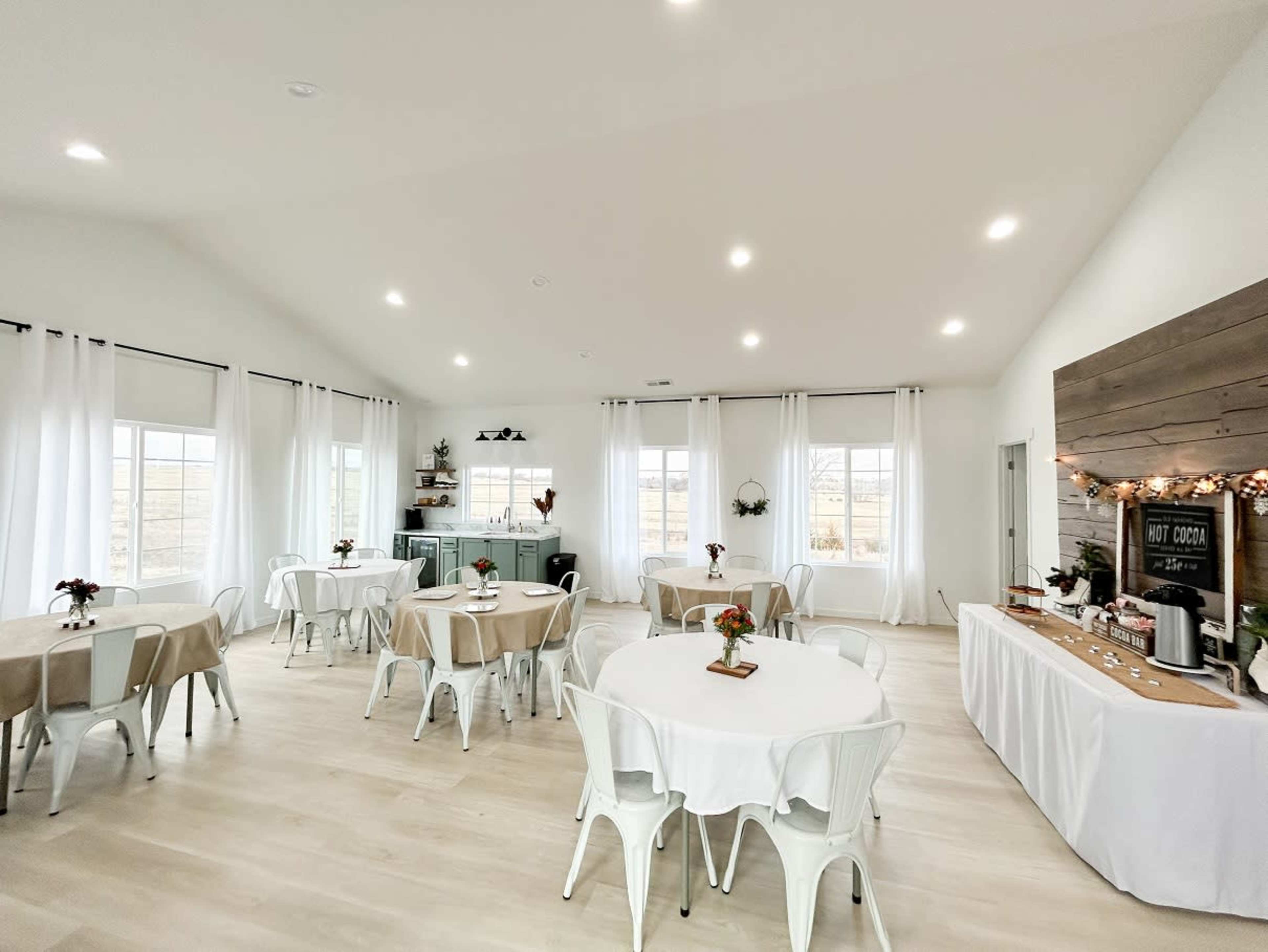 A bright, airy event space with several round tables set for guests, large windows, and a decorated service area featuring a coffee station.