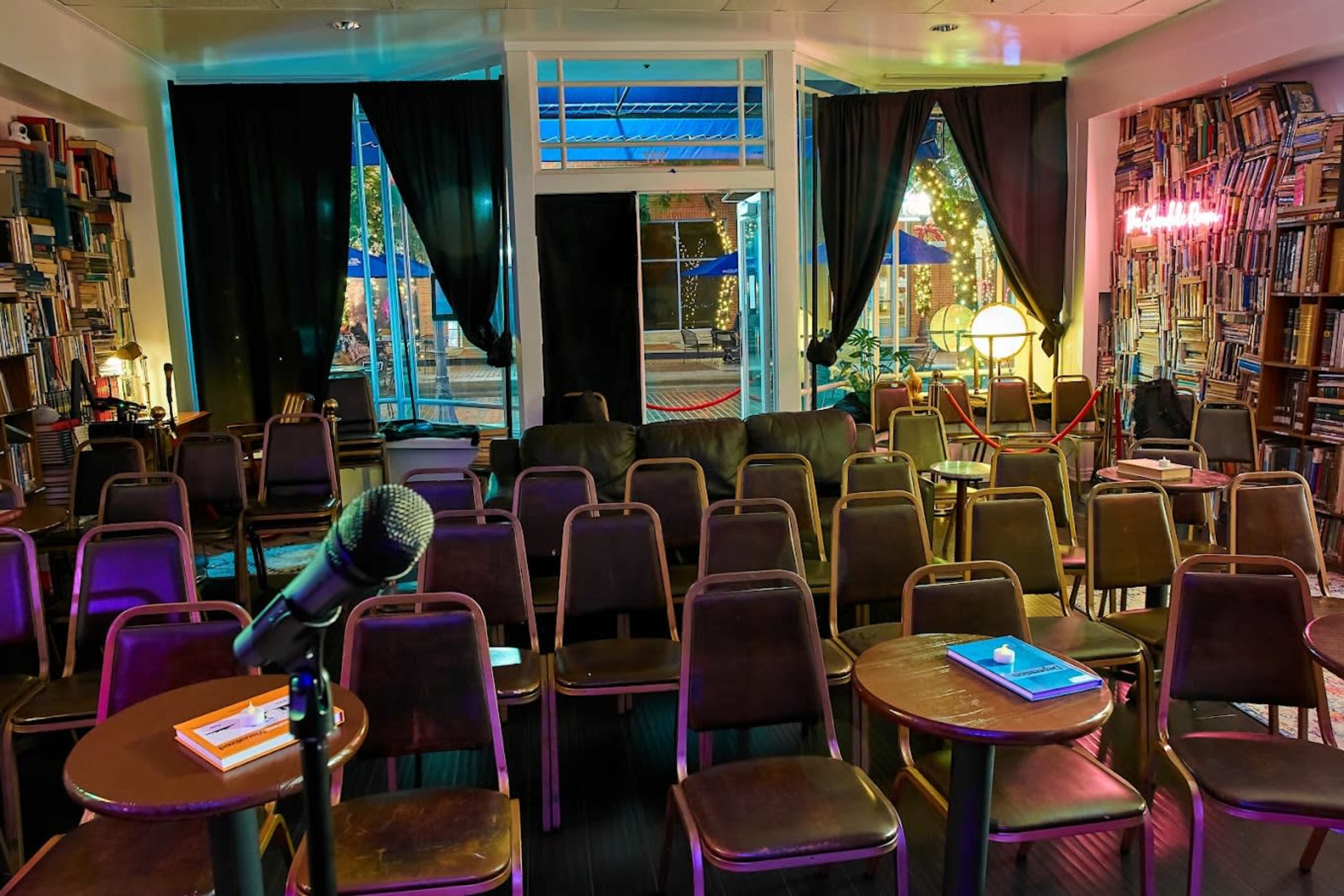 The image shows a cozy performance space with rows of chairs facing a small stage, surrounded by bookshelves and warm lighting.