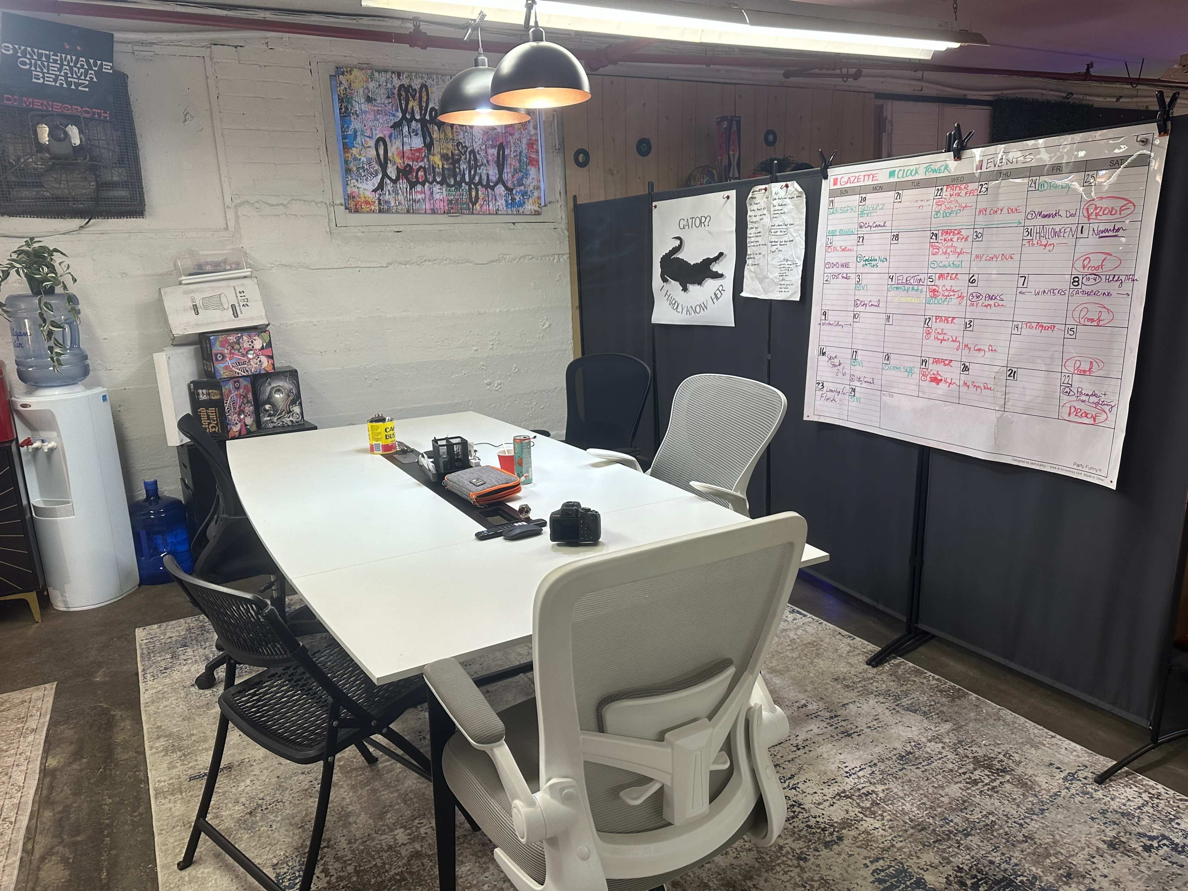 The image shows a modern meeting room with a large white table surrounded by chairs, a whiteboard filled with notes, and various items like water bottles and a plant in the background.