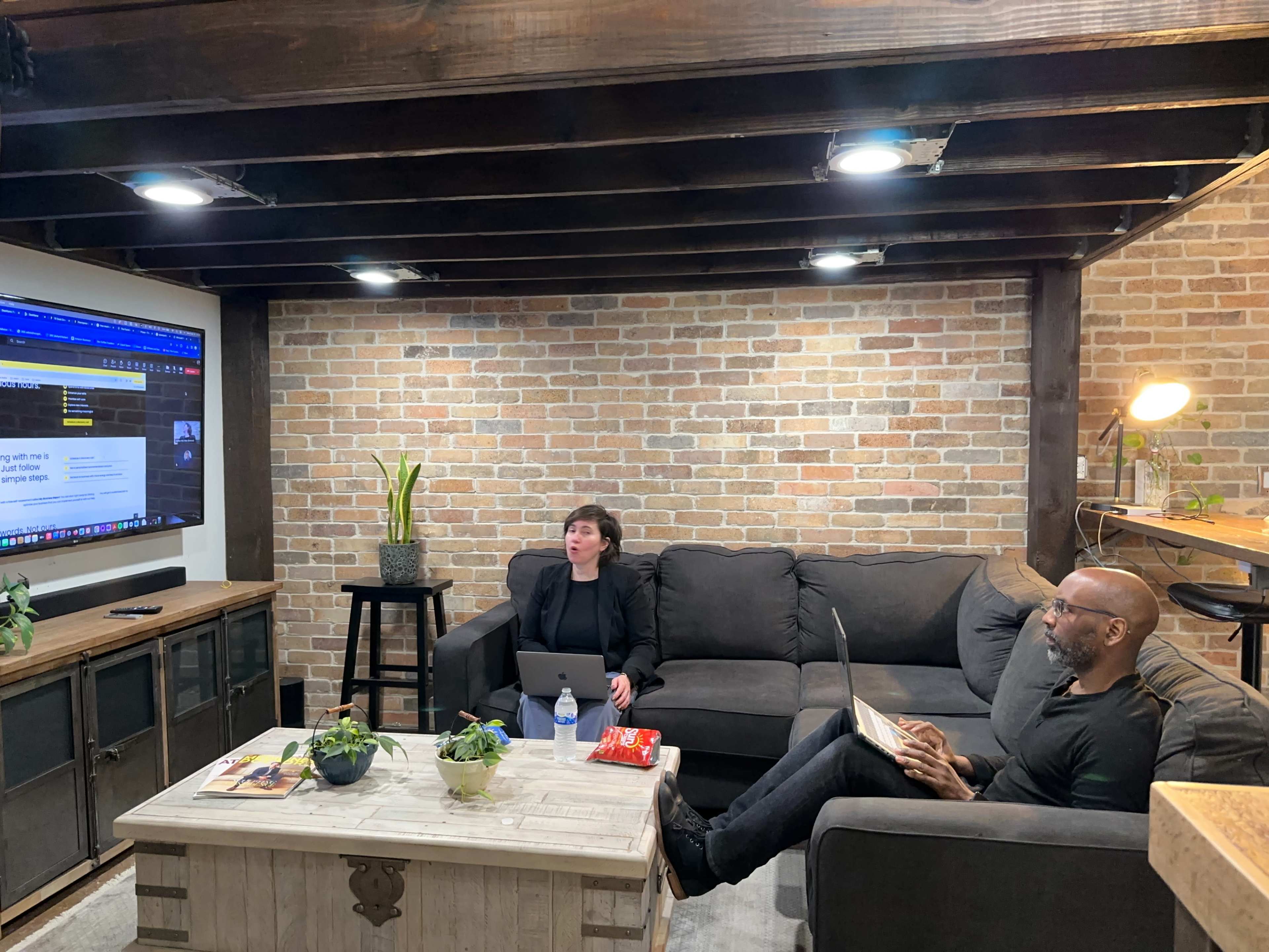 Two people are seated in a cozy room with a brick wall, working on laptops while a large screen displays information in the background.