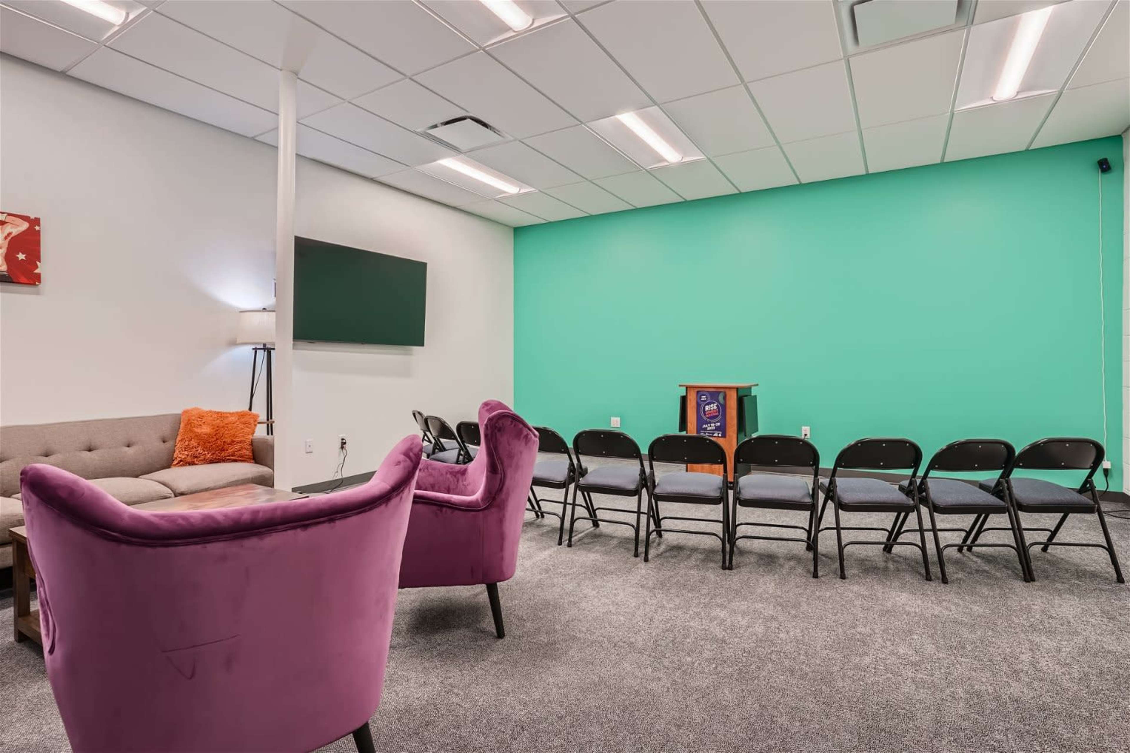 A room with a green wall, a sofa, and several black chairs arranged in a semicircle facing a small wooden podium.