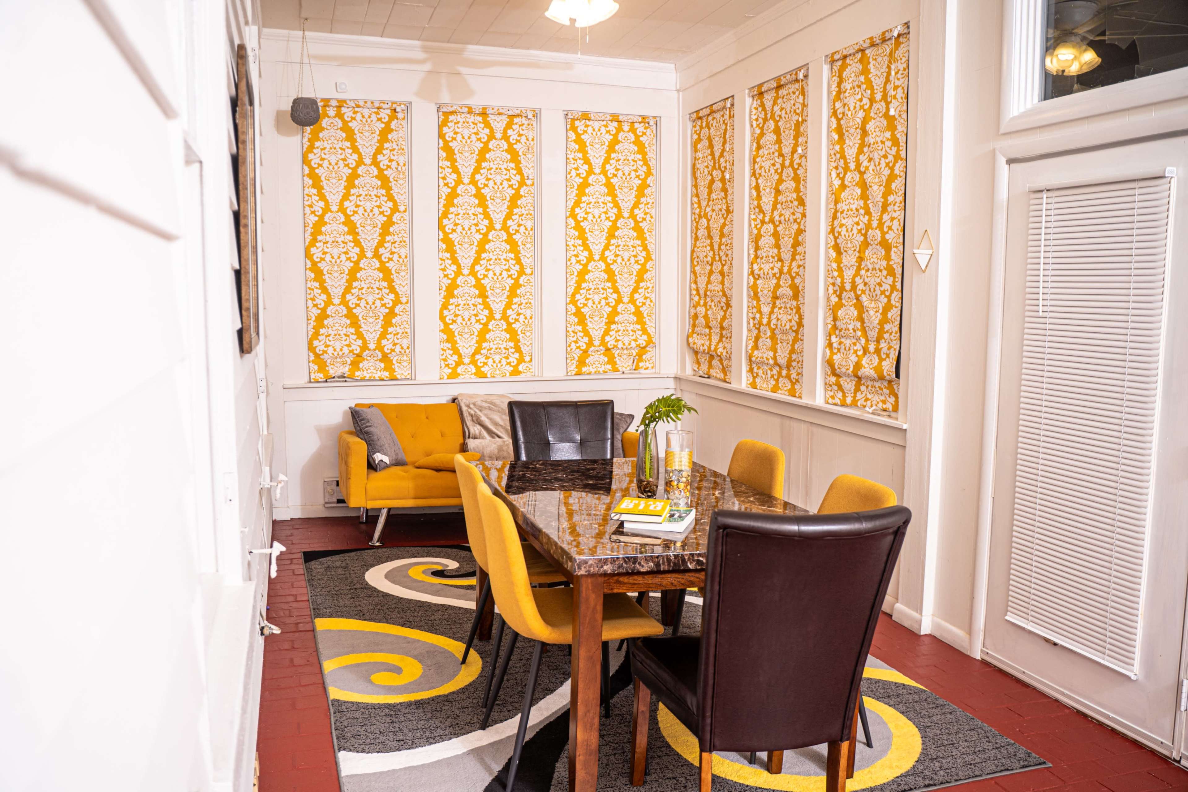 A dining area features a table surrounded by several chairs, with bright patterned curtains and a yellow accent chair adding color to the space.
