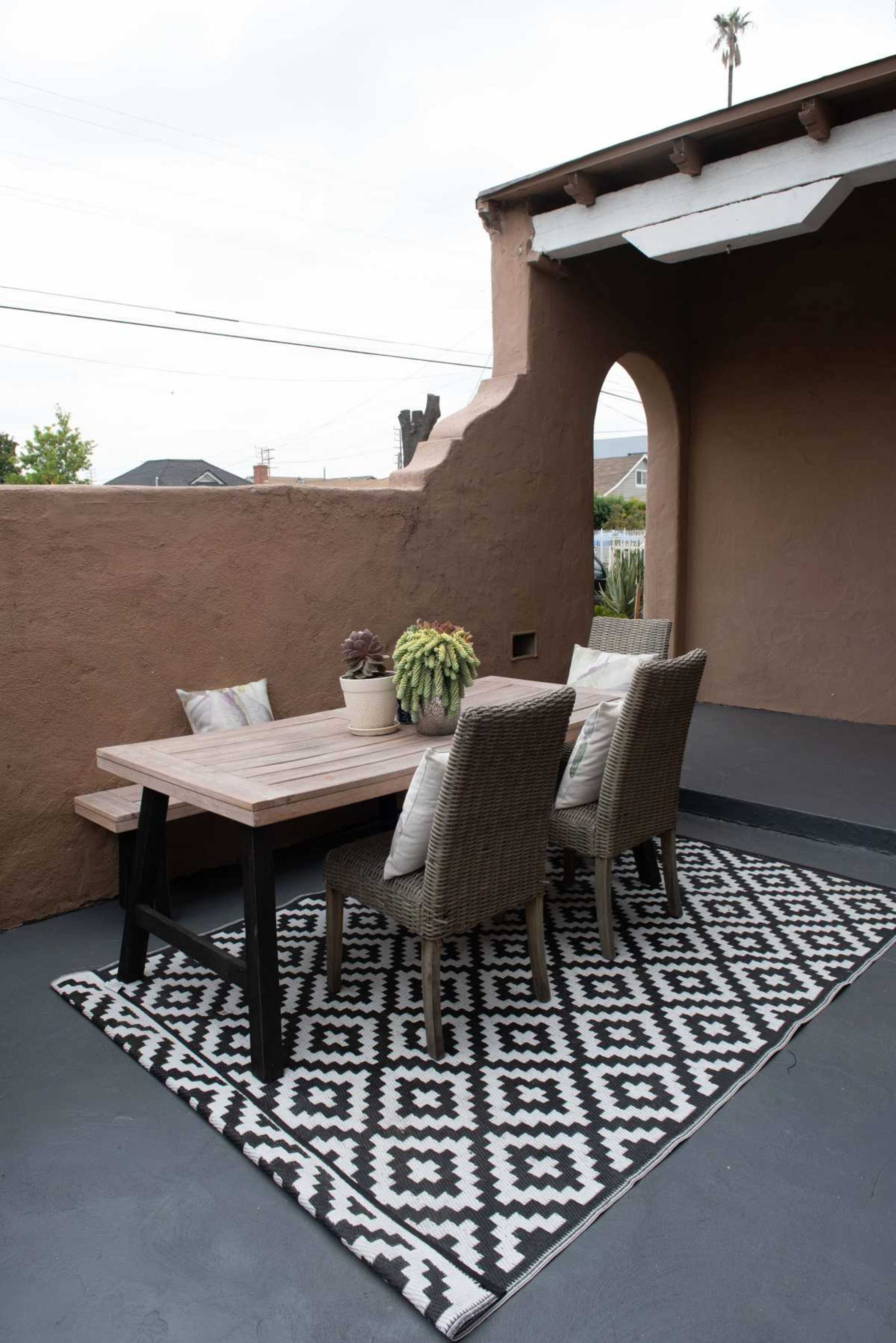 A wooden table with chairs is set on a patterned rug in an outdoor patio area surrounded by a decorative wall.