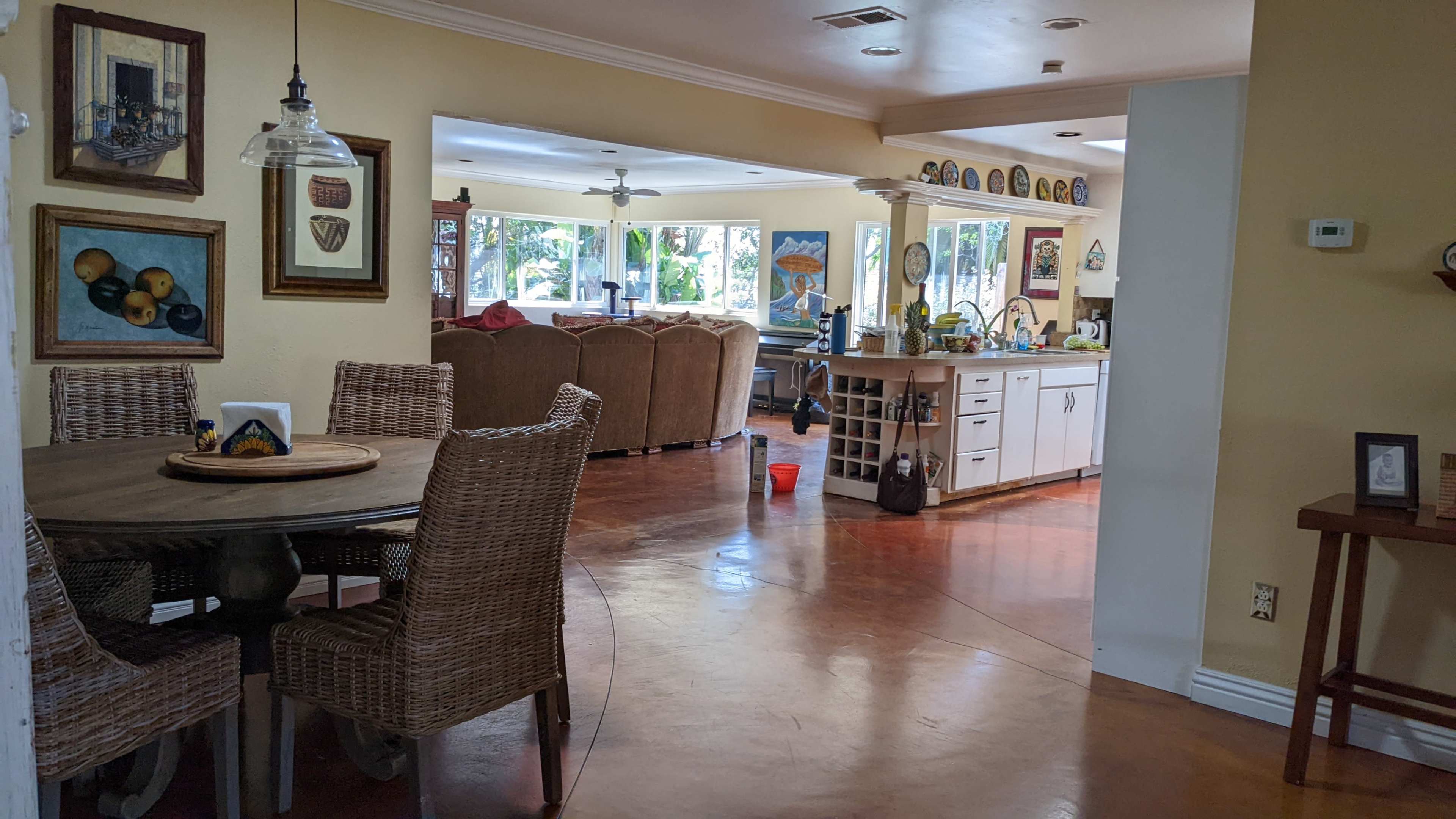 The image shows an open-concept living space with a dining area featuring a round table and woven chairs, leading to a kitchen and cozy seating area.