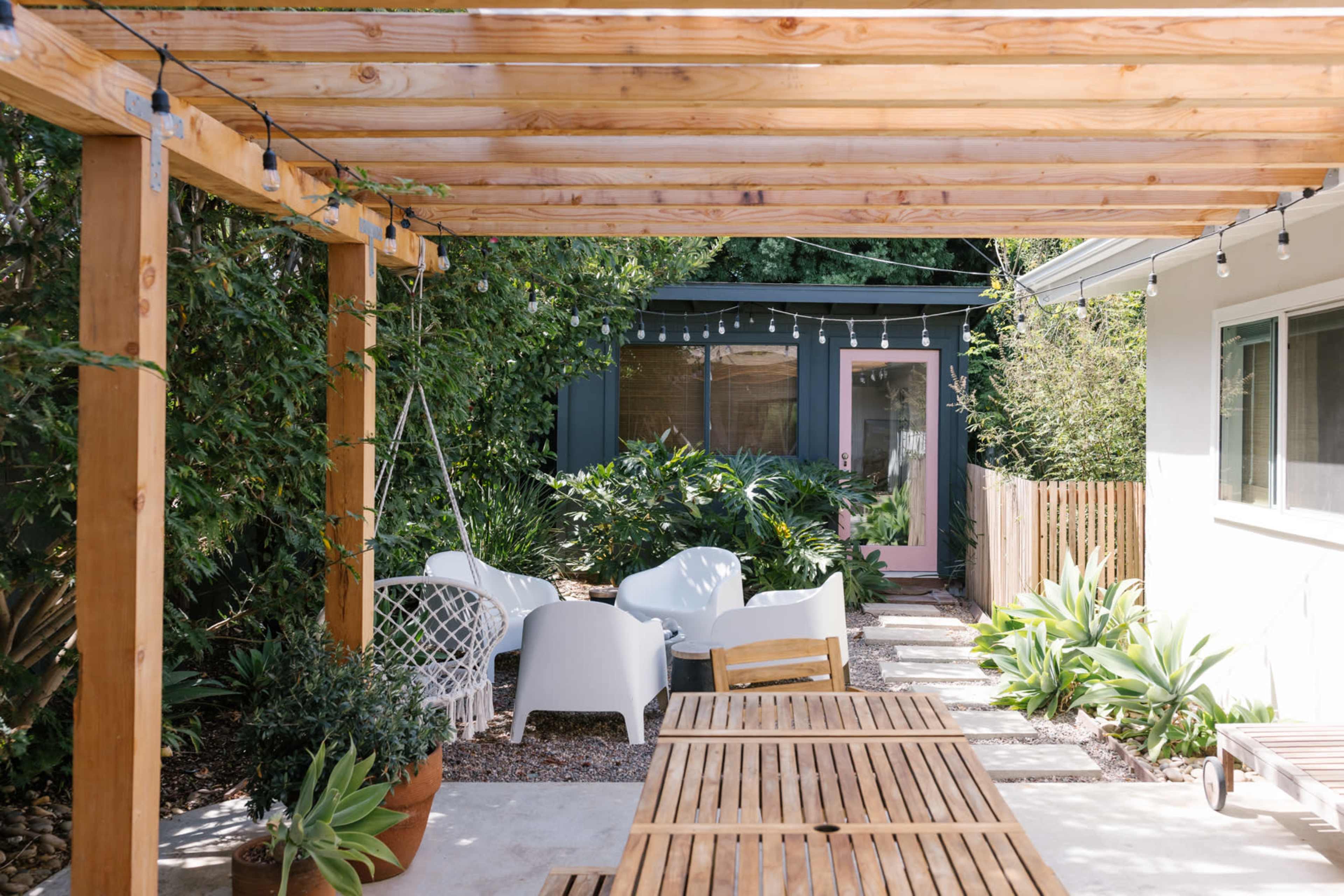 The image shows a backyard area featuring a wooden pergola, outdoor seating with white chairs, a table, and lush greenery in the background.