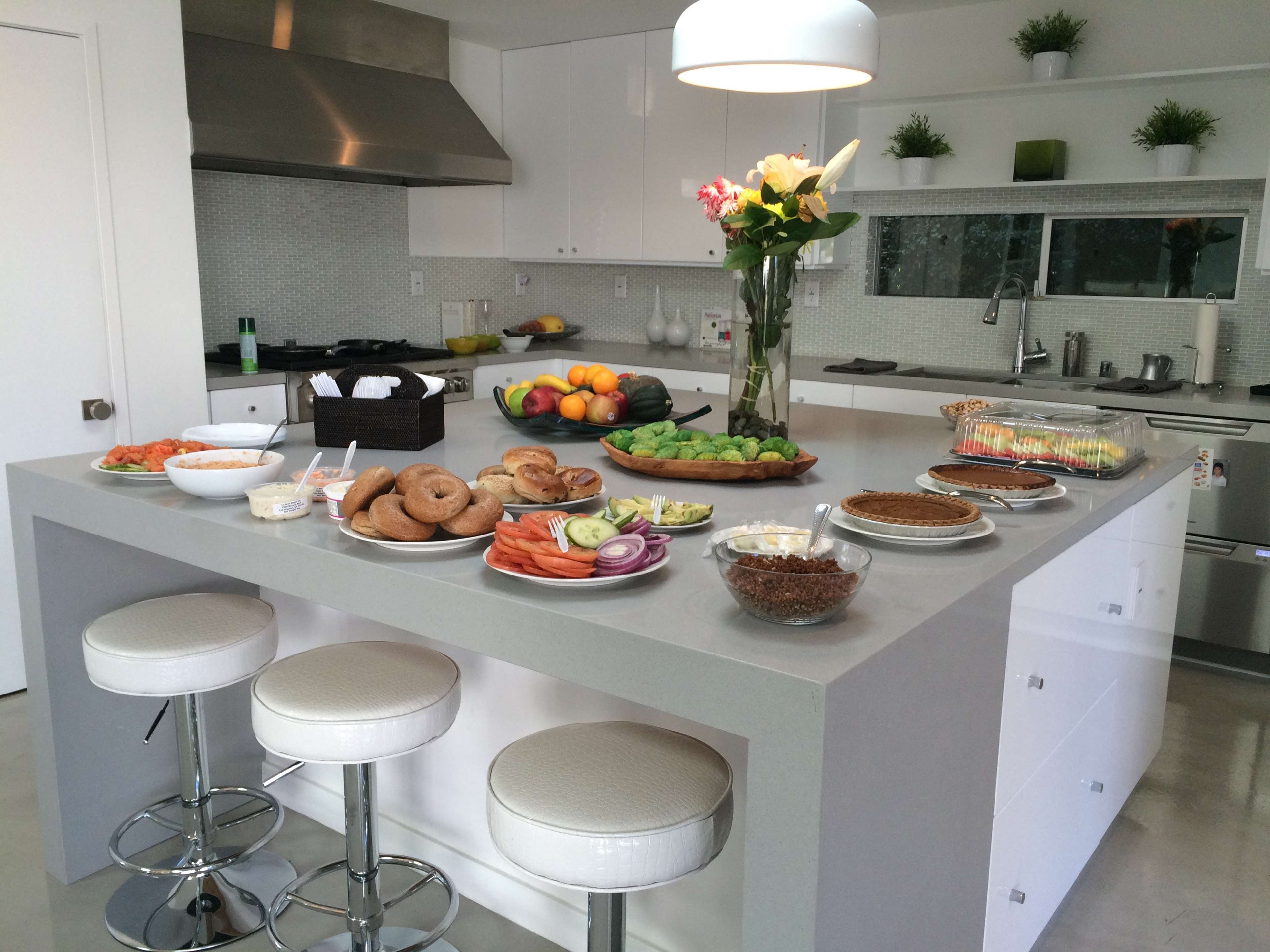A modern kitchen island is set with various dishes, including bagels, vegetables, fruits, and desserts, surrounded by sleek white cabinetry.