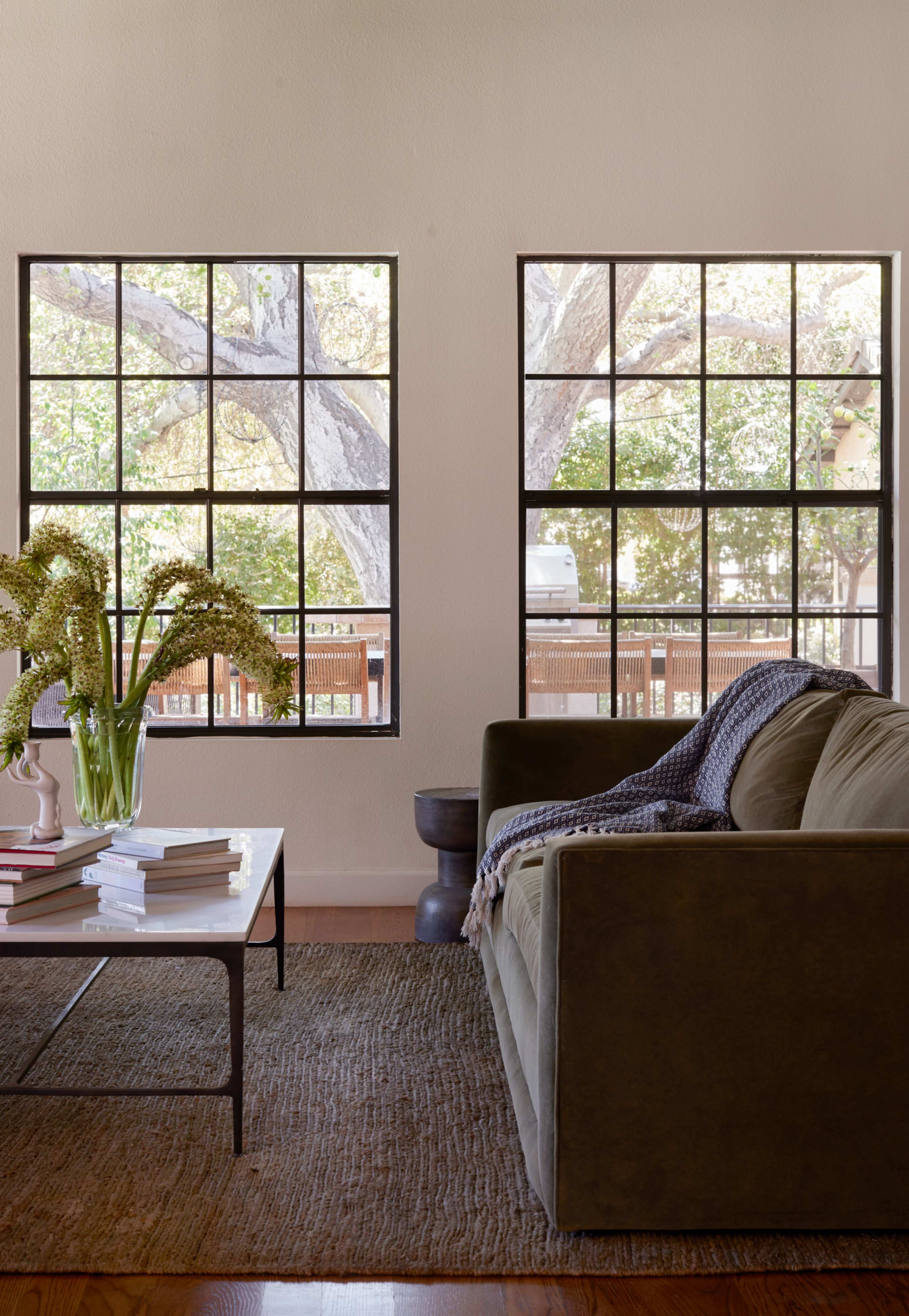 A cozy living room features a green sofa, a coffee table with books, and large windows offering a view of trees outside.