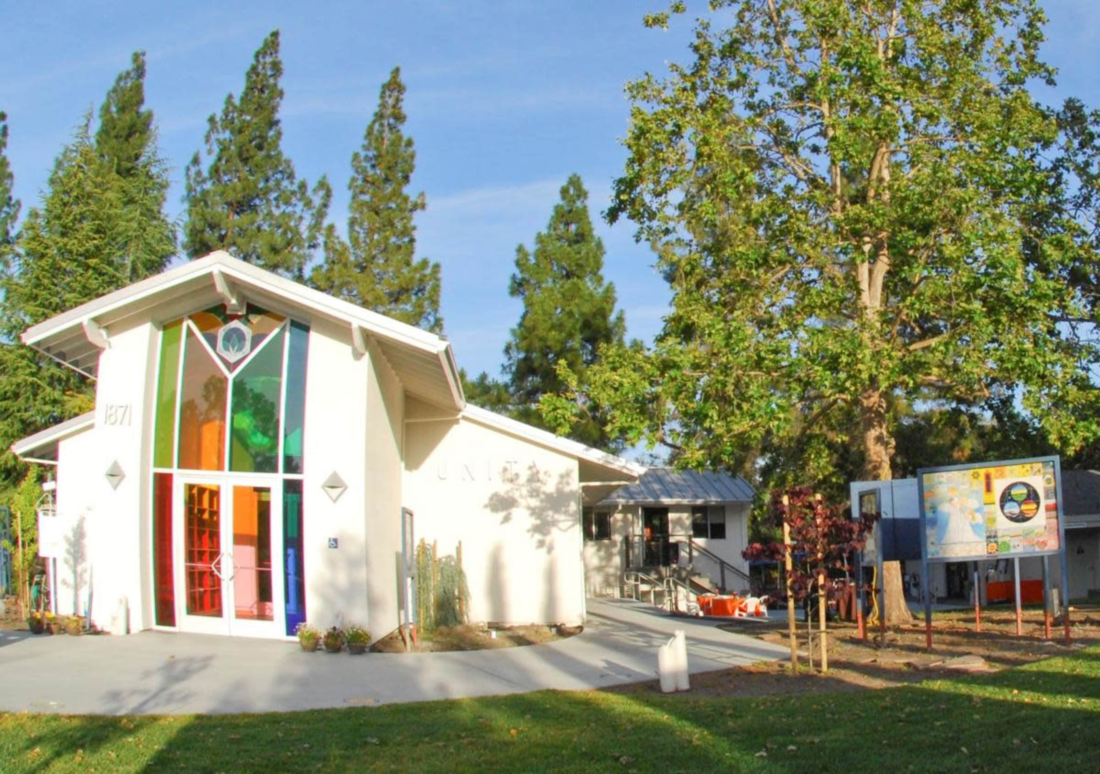 A modern, single-story building with a colorful glass entrance surrounded by greenery and a large tree.